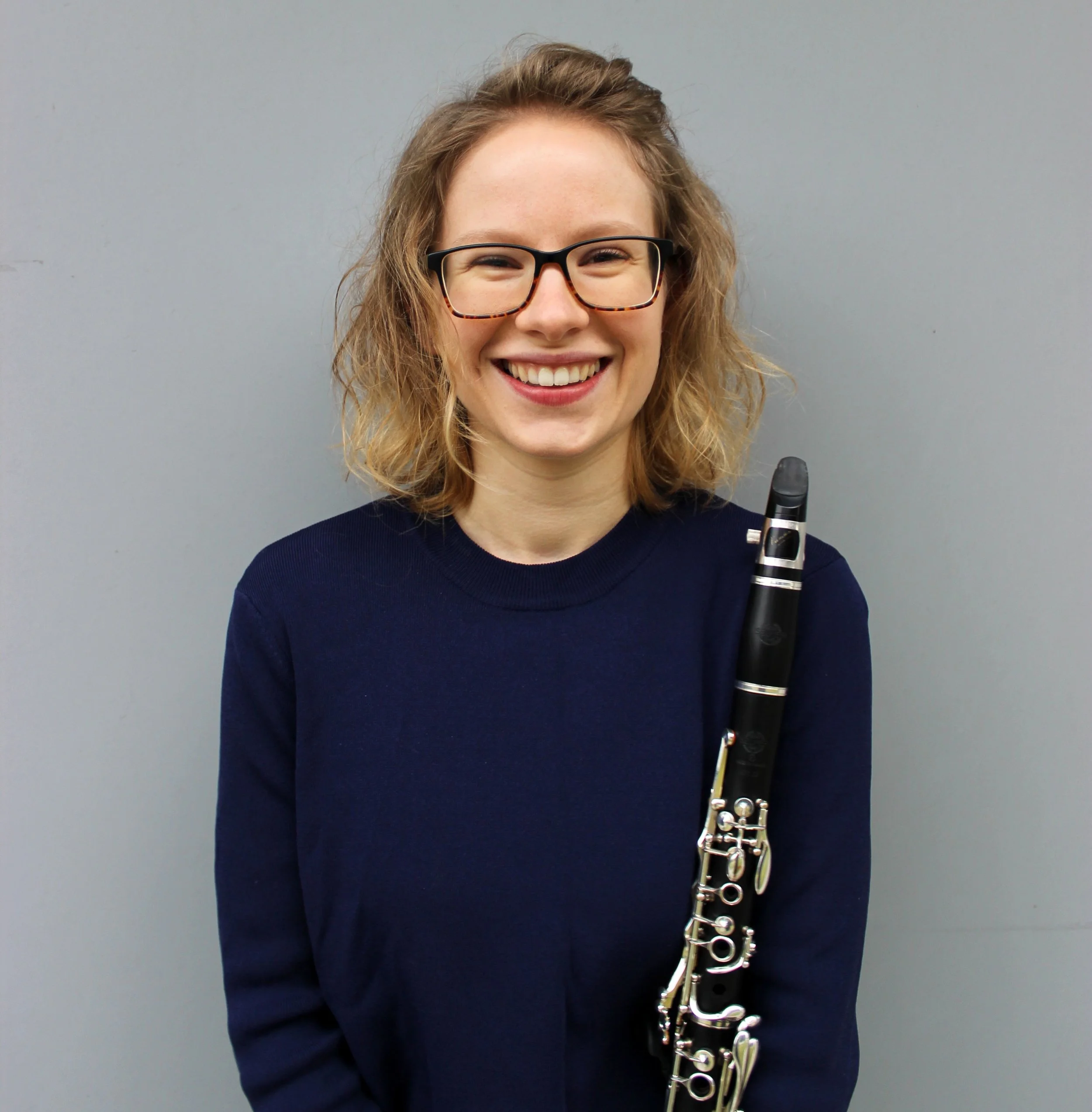 A woman with blonde hair, wearing glasses and a navy blue sweater, smiling and holding a clarinet against a plain grey wall.