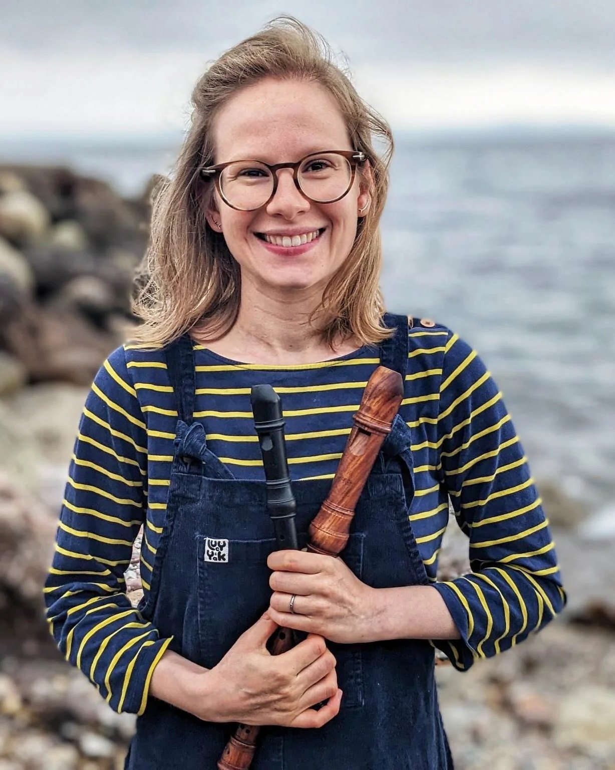 A woman with glasses, holding recorders, standing on a rocky shoreline with water and a cloudy sky in the background, smiling at the camera.