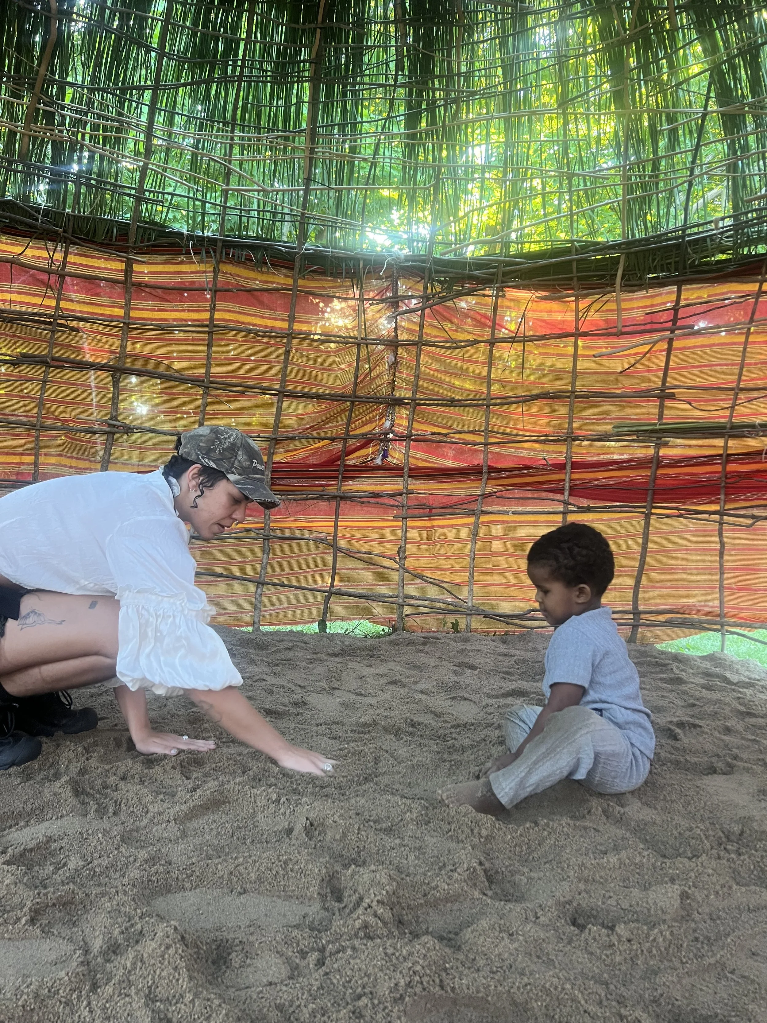 A person with curly hair and a white shirt crouching and helping a young boy in gray clothes dig in the sand inside a shaded outdoor area with bamboo fencing and greenery outside.