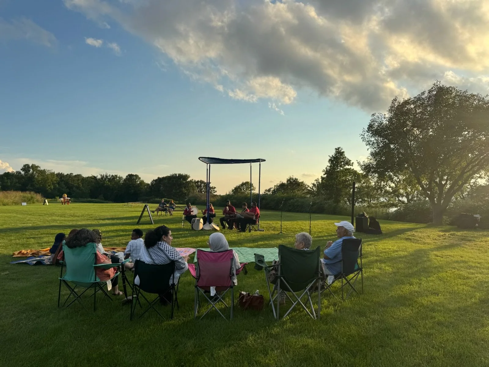 Outdoor concert in a park with people seated on chairs and blankets watching musicians perform on a small stage as the sun sets.