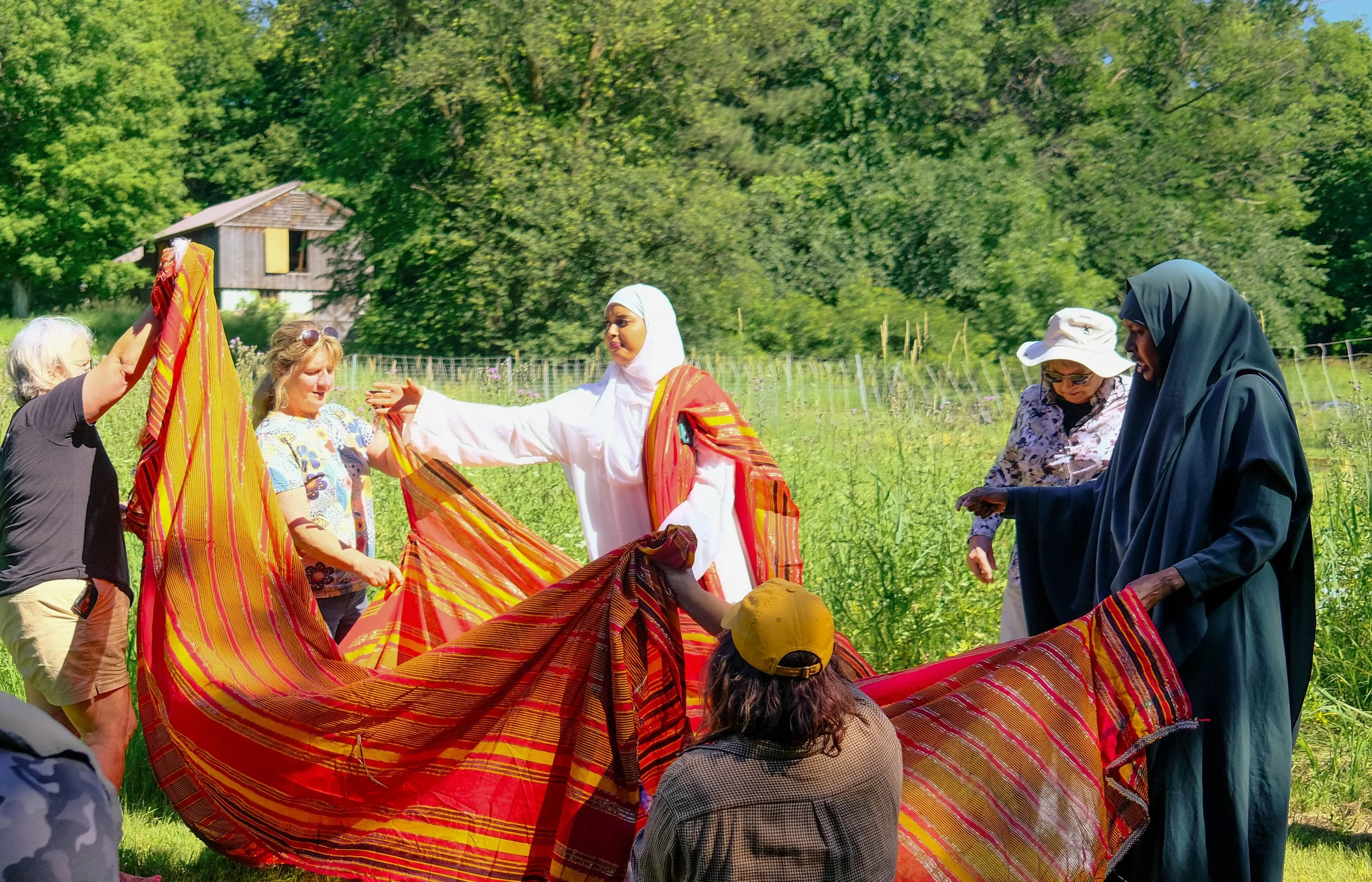 Group of diverse women and children outdoors, holding a brightly colored striped fabric, with a lush green landscape and small wooden house in the background.