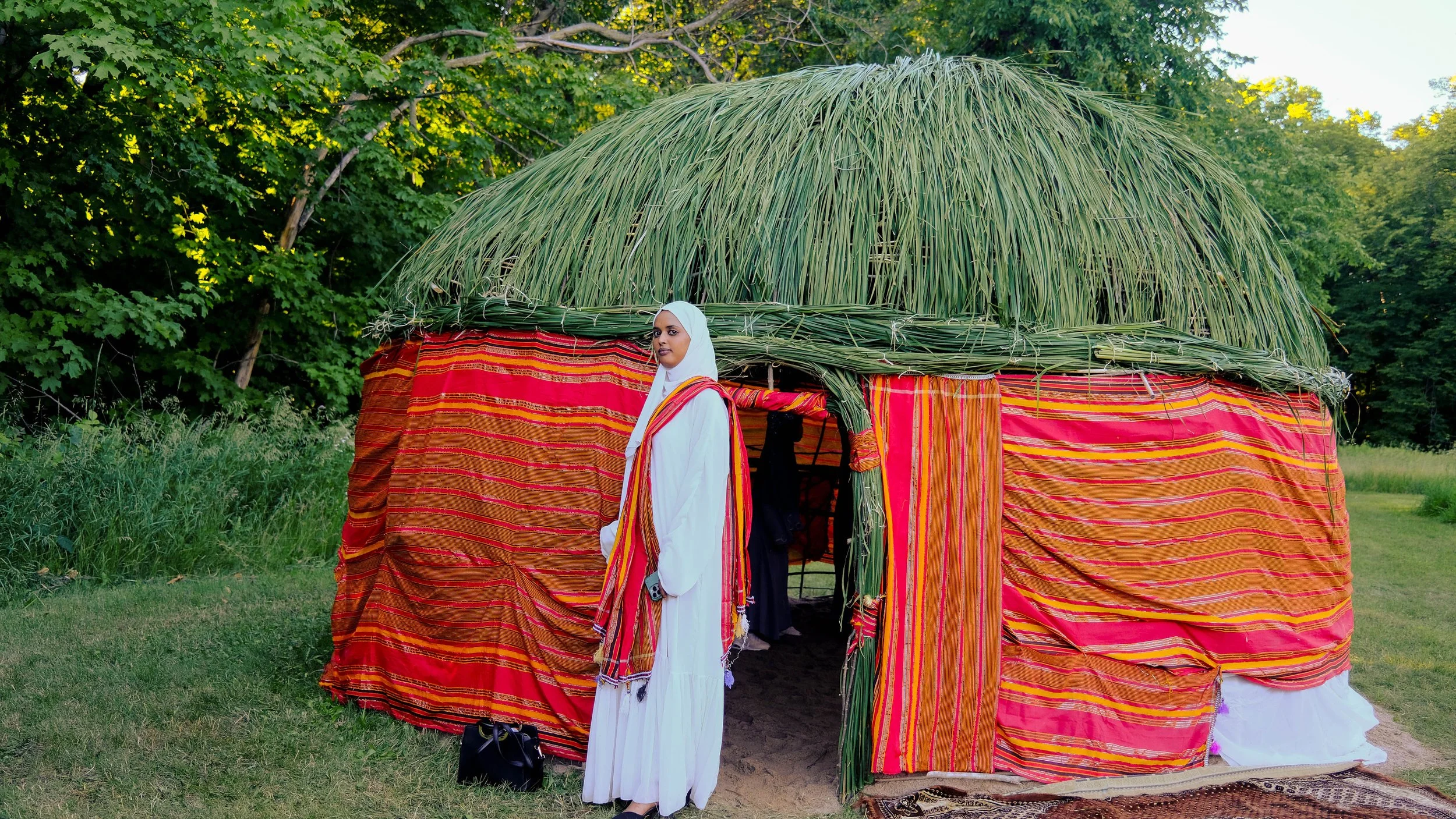 A woman dressed in white standing in front of a traditional tent made of colorful striped fabric and green grass roofing, set outdoors surrounded by green trees and grass.