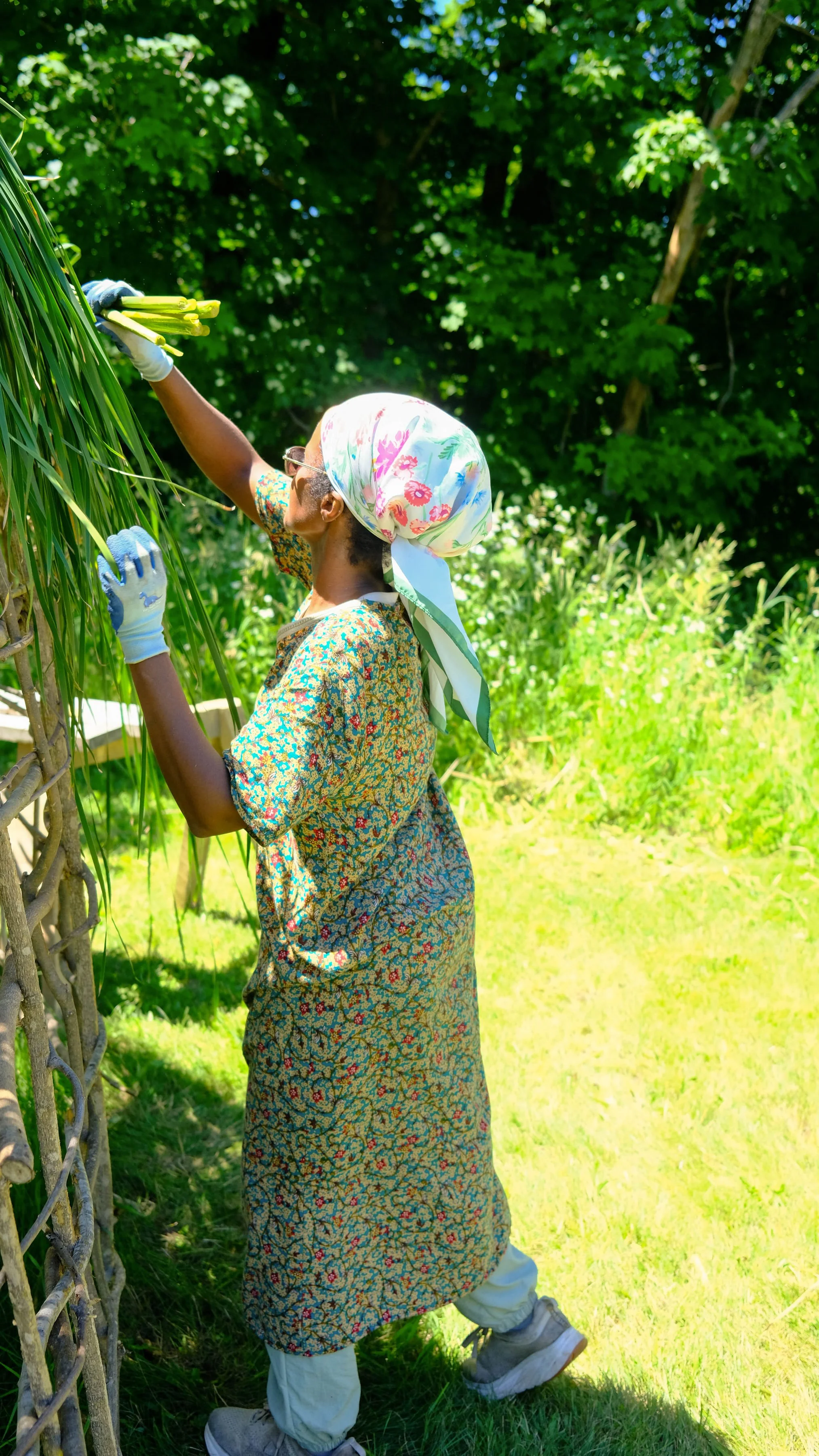 A person wearing a floral headscarf, glasses, a patterned dress, and boots, harvesting or tending to plants in a lush green outdoor garden.