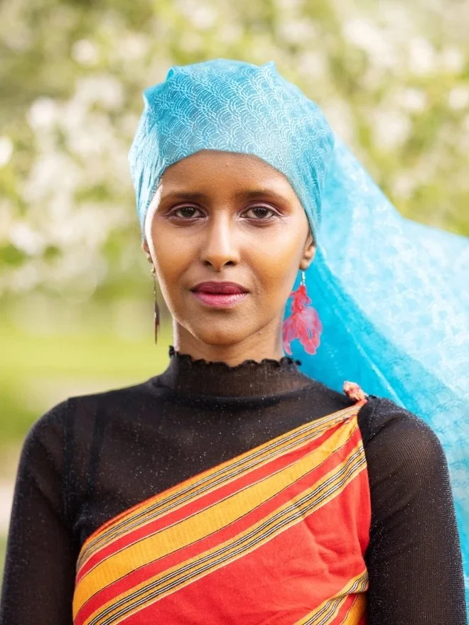 A woman wearing a black top, a traditional red and yellow saree, a blue headscarf, and red earrings, standing outdoors with a blurry green background.