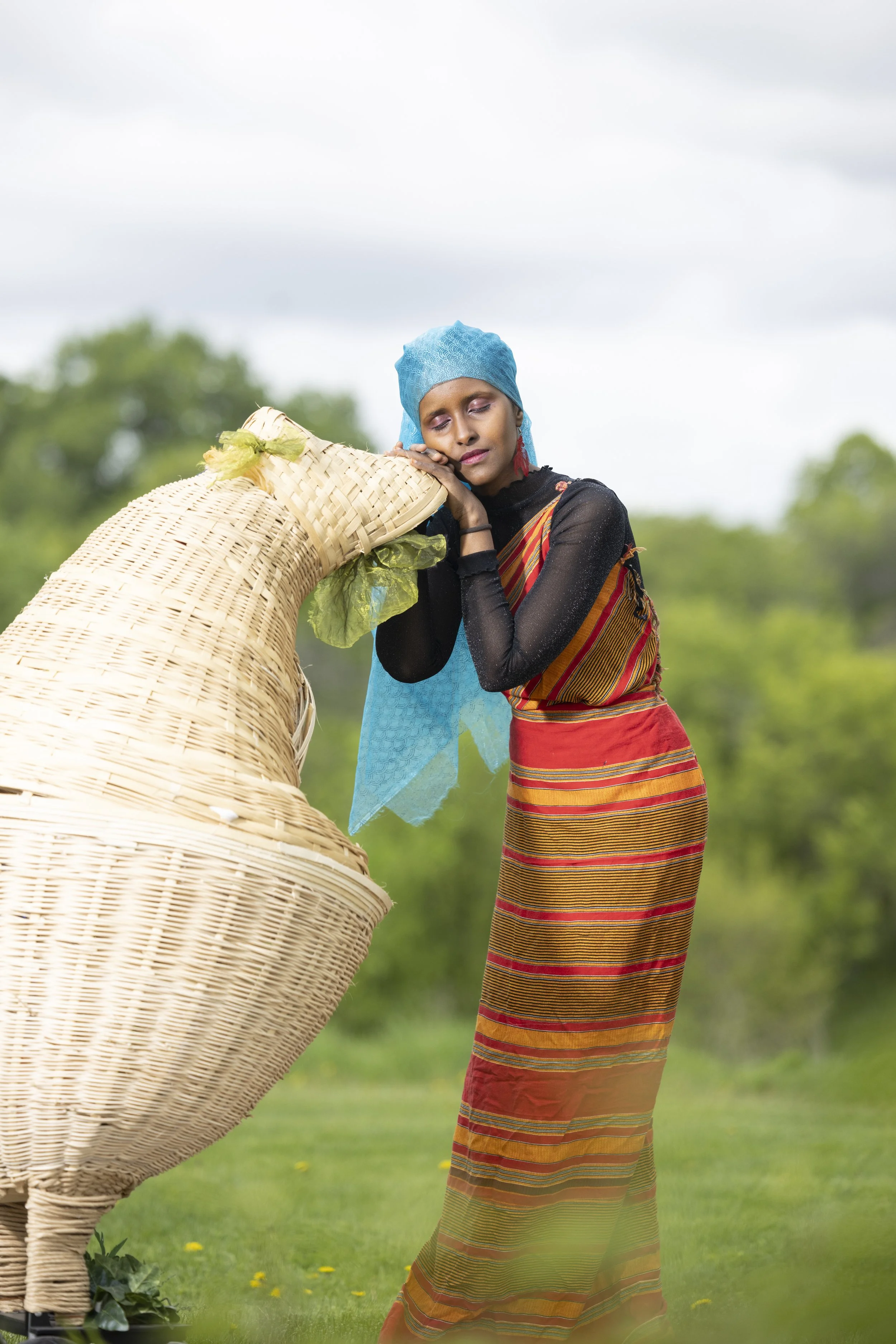 A woman in traditional attire resting her head on a large woven basket outdoors with a green field and trees in the background.