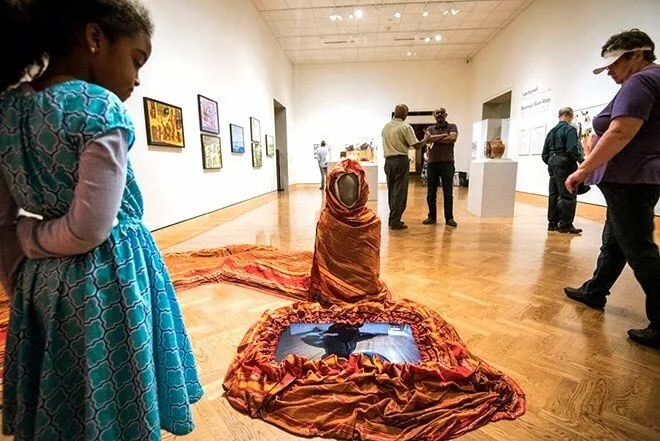 A girl in a blue dress looks at an art installation in a gallery, consisting of a fabric-wrapped object with a hat on top and a reflective surface, surrounded by other visitors and artwork on the walls.