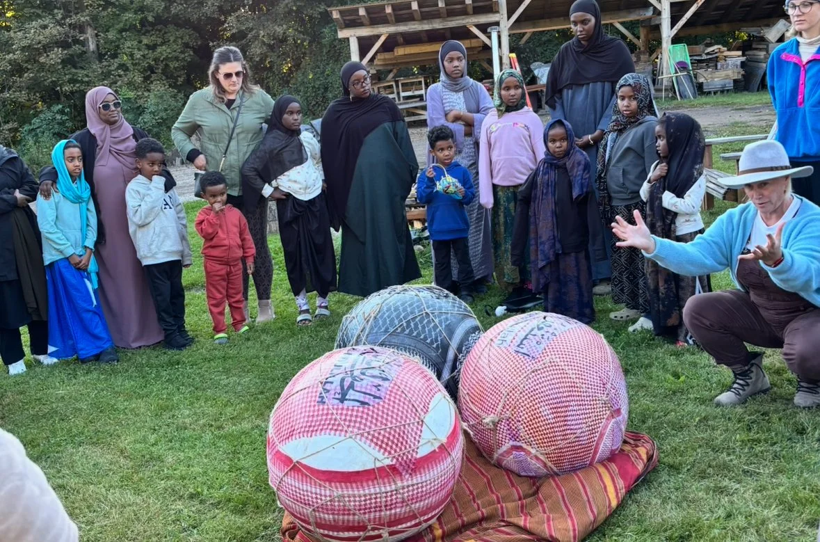 A group of women and children gathered outdoors around a person who is speaking and gesturing with their hands. There are large, wrapped objects on a cloth on the grass in front of them.