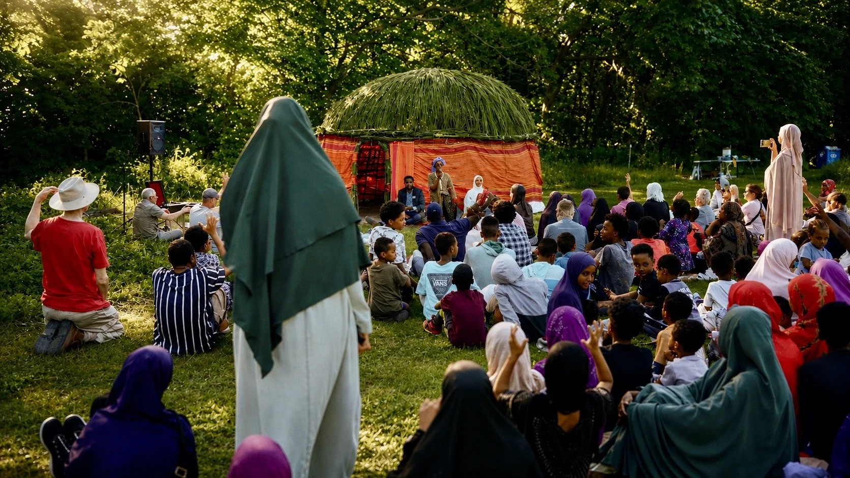 A diverse group of people gathered outdoors on a grassy area, attending a cultural or community event with a stage covered by a green and orange tent, where a speaker is addressing the audience. The crowd includes children and adults, some wearing tr