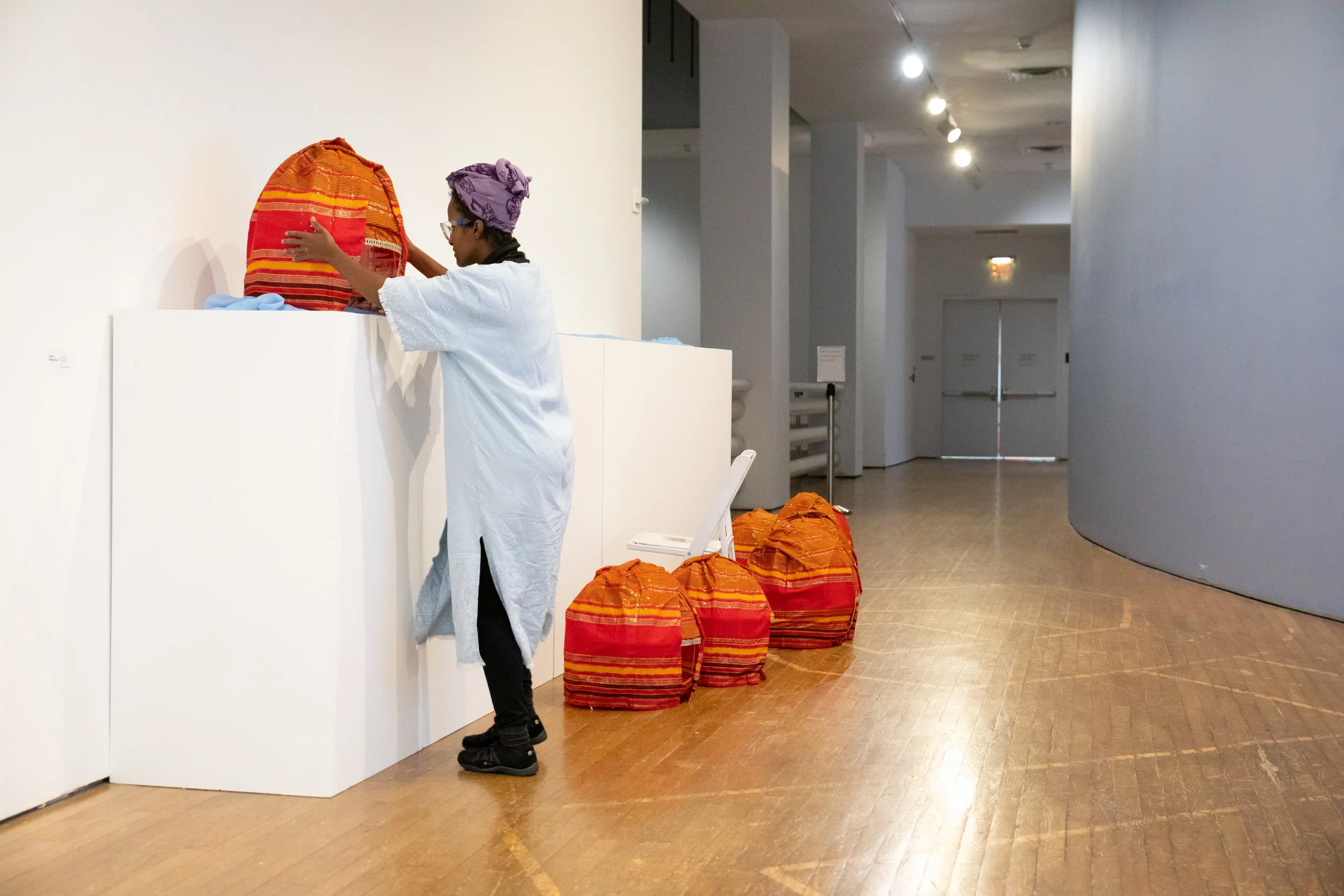 A person wearing a purple headscarf, glasses, and a white gown is placing a striped red and orange bag on a white display counter in an art gallery or museum. Multiple similar bags are scattered on the floor nearby. The space has light-colored wooden