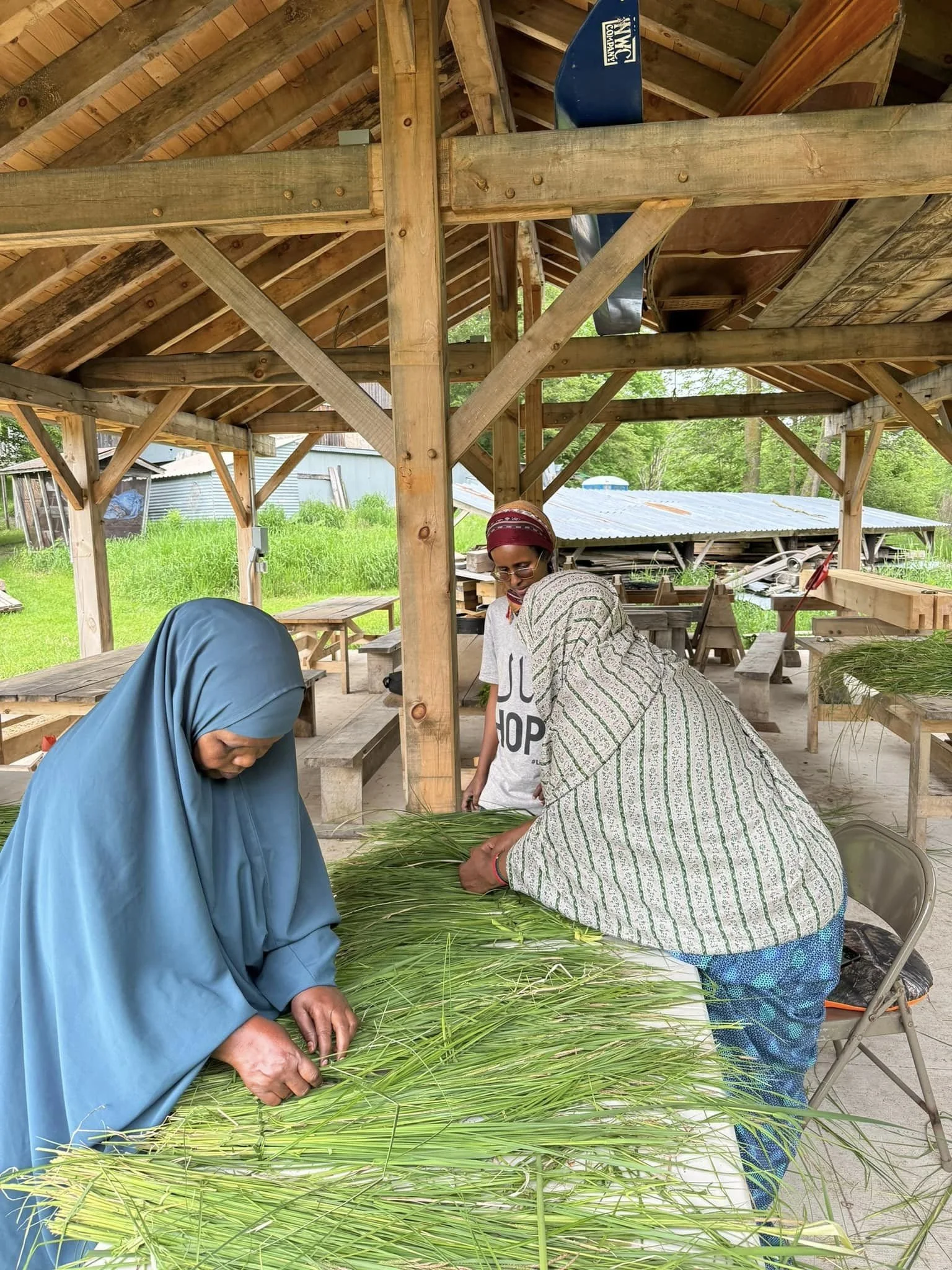 Three women are working with fresh green grass in a rustic wooden outdoor shed with a sloped roof, surrounded by lush greenery and trees.