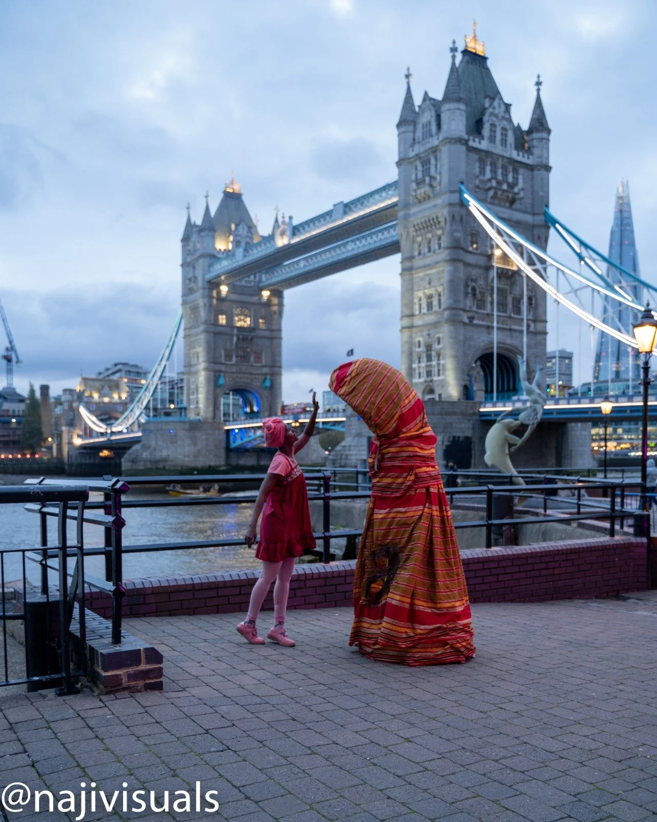 A woman wearing a colorful traditional African dress and head wrap is giving a high five to a young girl in a pink dress and headwrap by the Thames River with Tower Bridge illuminated in the background in London, England.