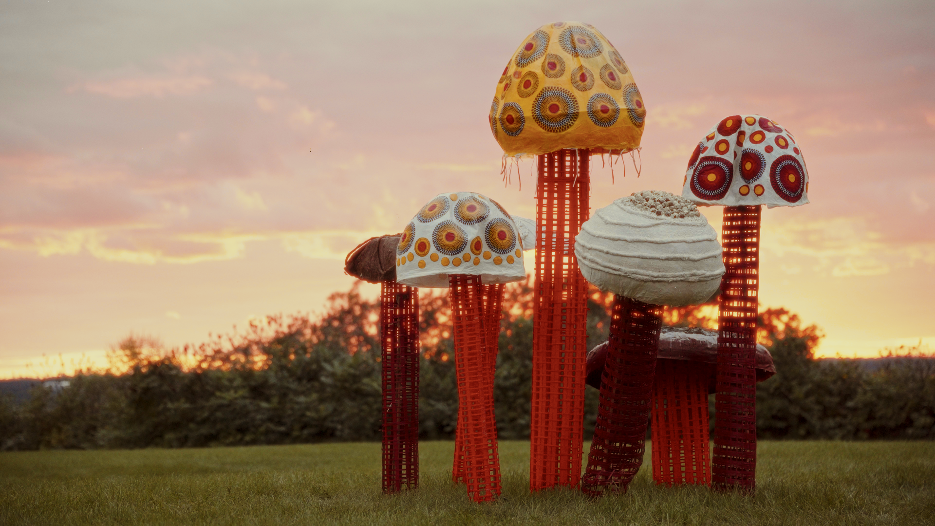 Art installation of giant mushroom sculptures with colorful patterned caps and red lattice stems, set against a sunset sky with trees in the background.