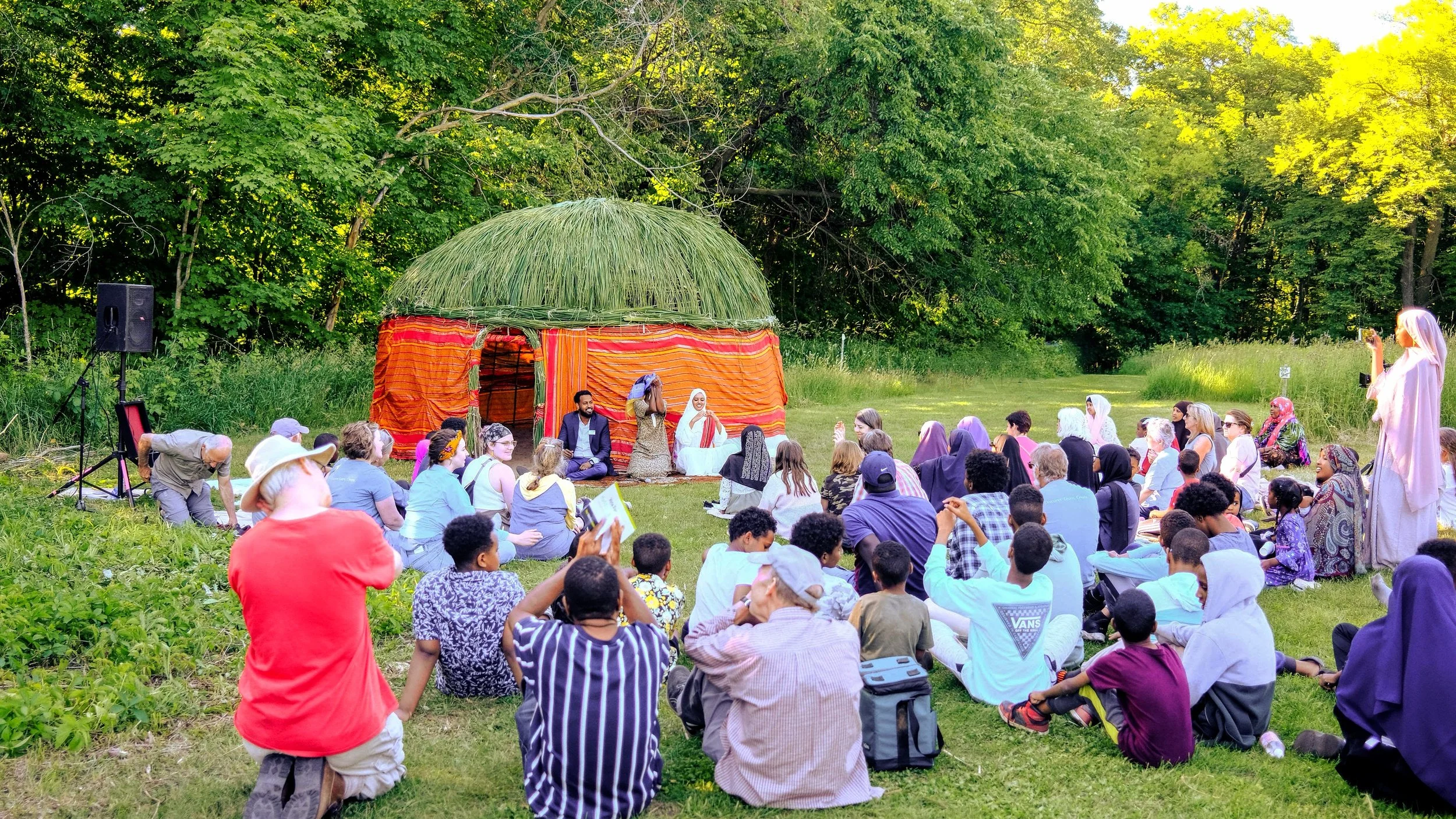 A diverse group of people gathered outdoors in a grassy area surrounded by trees, watching a cultural performance with a traditional straw hut backdrop. The scene includes individuals capturing the event on their phones, sitting on the grass, or knee