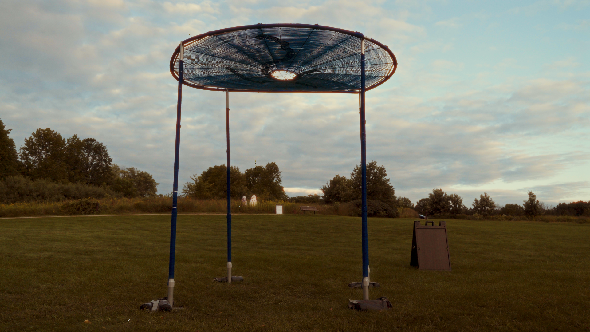 A large outdoor solar cooker with three tall blue poles supporting a circular reflective surface at the top; the sun is shining through the opening in the reflector. The scene is set in a grassy park with trees and a cloudy sky in the background.