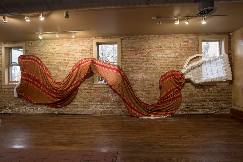 Indoor art installation resembling a sofa, with a striped fabric draped over a brick wall and a partially collapsed whitestructure on the right side.