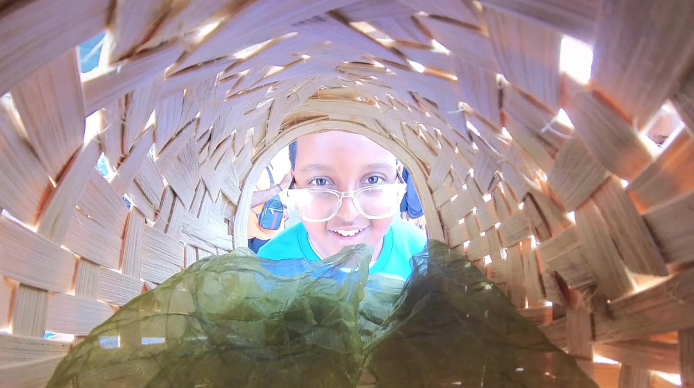A young person with glasses smiling inside a woven basket or tunnel, holding a large green leaf