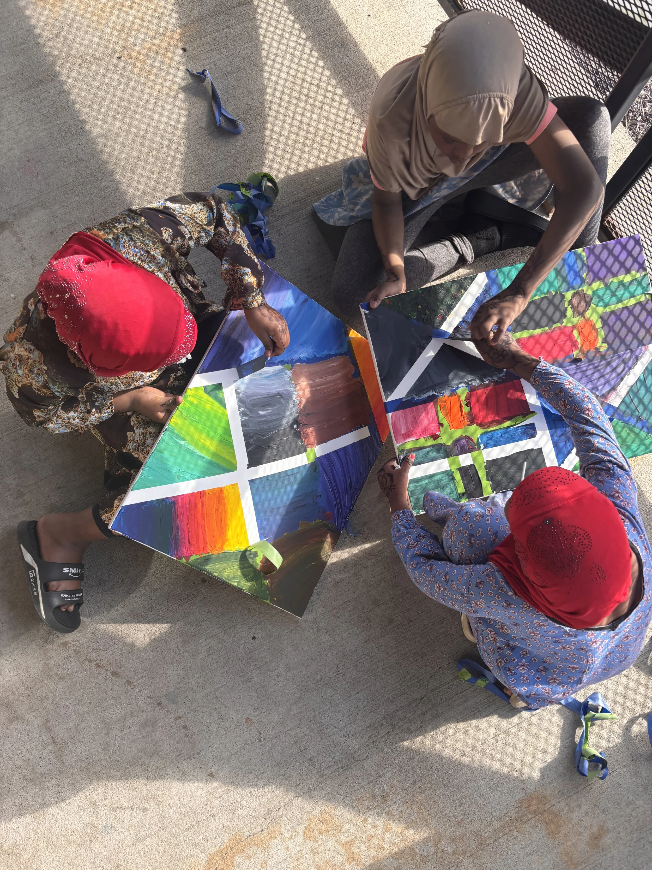 Two children dressed in colorful clothing and headscarves are sitting on the ground, engaged in painting colorful pictures on large sheets of paper. There are scattered straps and shoes on the ground nearby, and the scene is illuminated by sunlight, casting shadows on the ground.