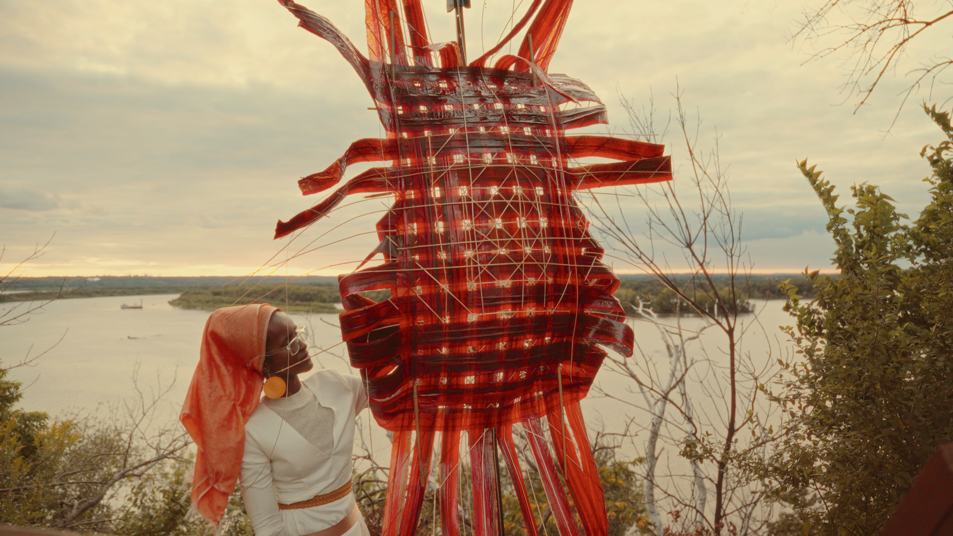 A woman wearing an orange headscarf and earrings holds a large, elaborate red and orange woven sculpture outdoors by a river, with trees and a cloudy sky in the background.