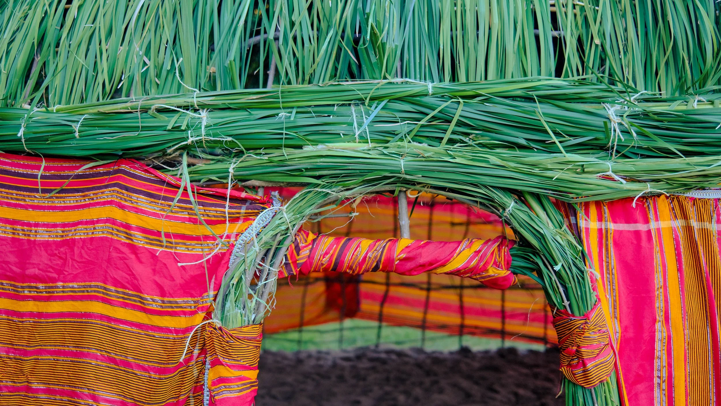 Colorful structure made of green grass or reeds, wrapped with ties, covered with vibrant red and orange striped fabric.