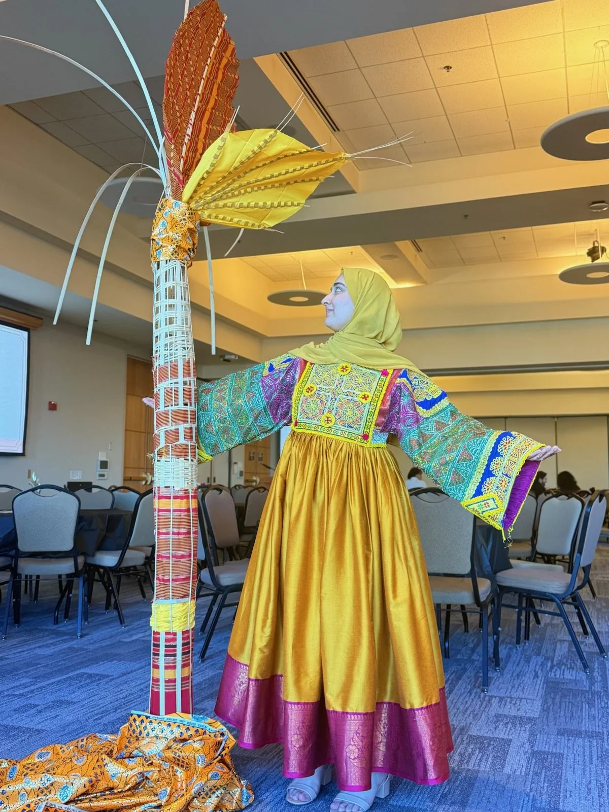 A woman in colorful traditional dress with a yellow skirt and embroidered top poses next to a large, colorful feathered sculpture at an indoor event. Chairs and a projection screen are visible in the background.