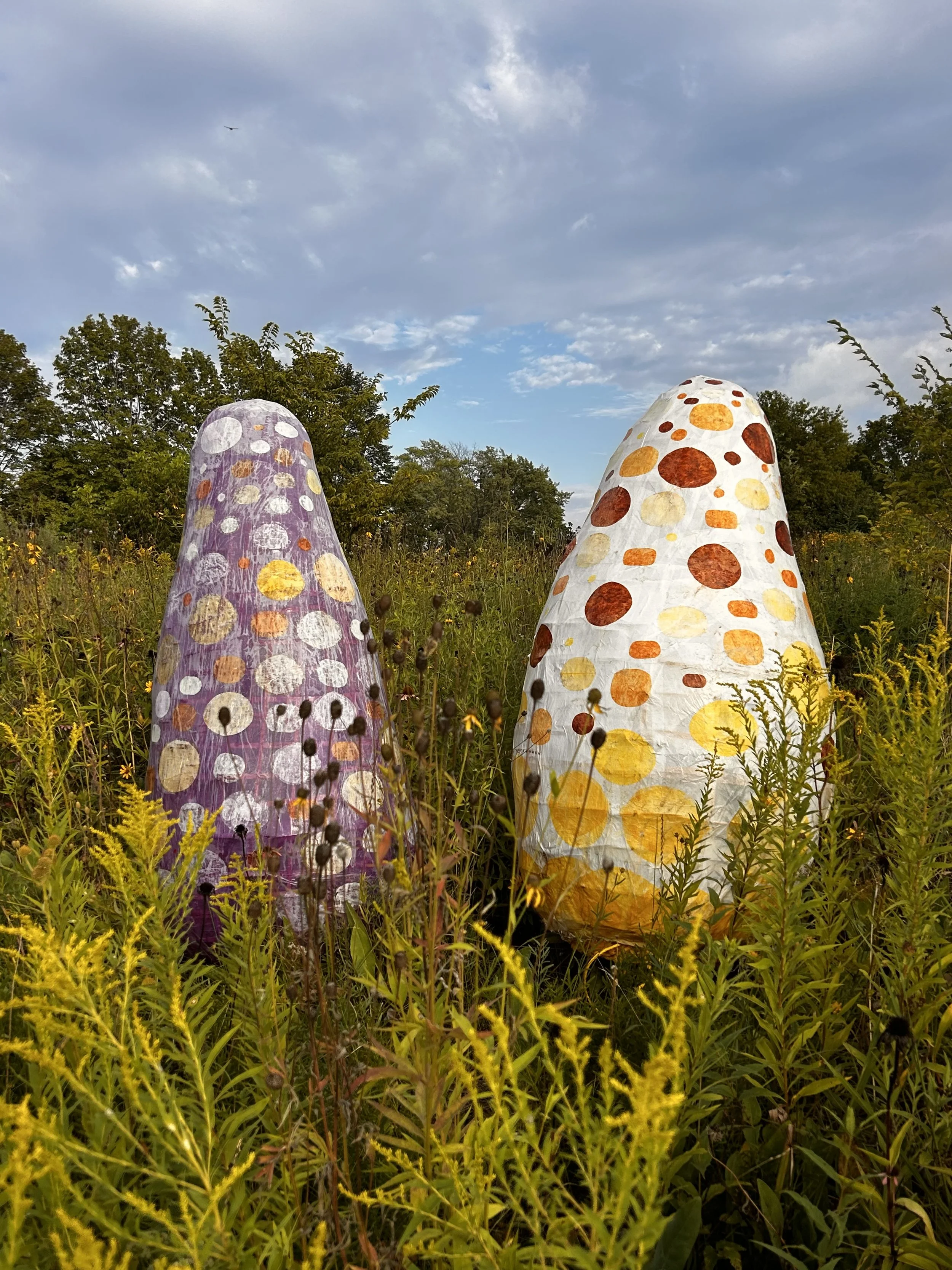 Two large, polka-dotted sculptures in a field of wildflowers with trees and a partly cloudy sky in the background.