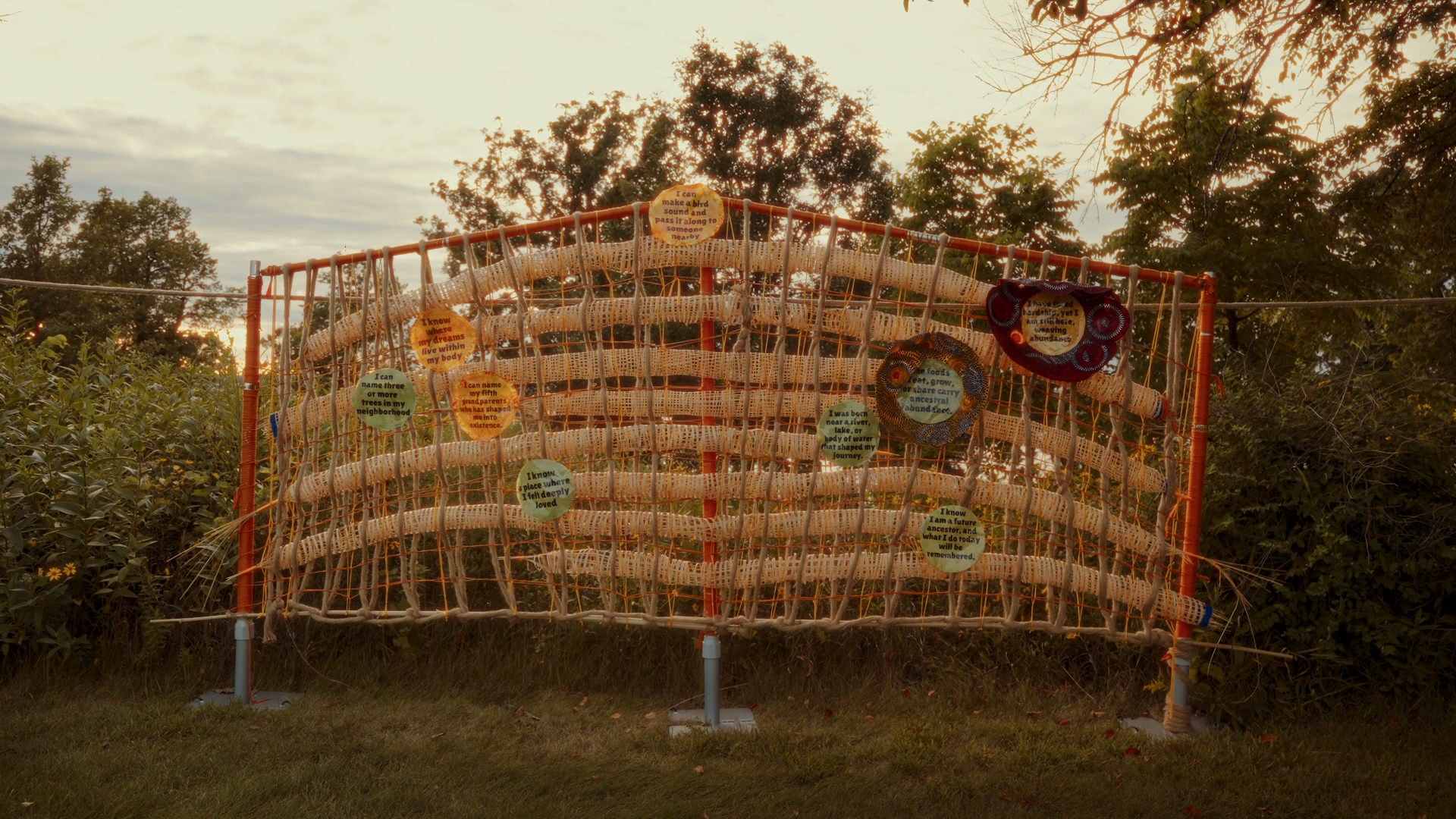 This is outdoor art installation featuring a large wall made of intertwined ropes and branches, decorated with colorful circular cutouts with handwritten texts about nature, trees, and personal memories, set against a backdrop of trees and a cloudy s