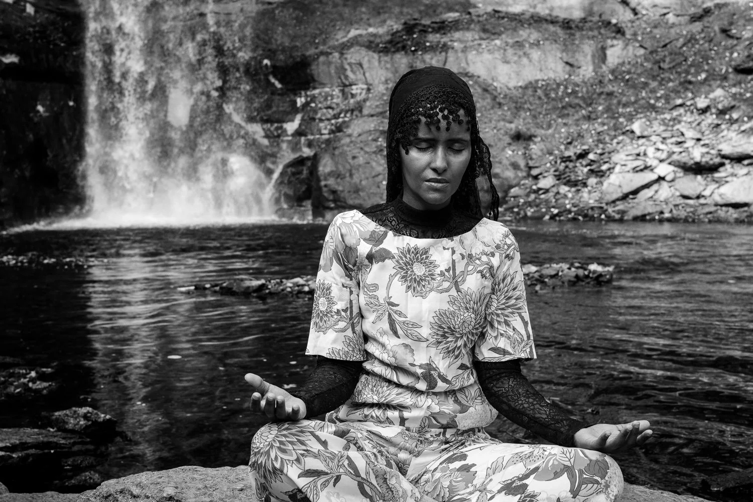 A woman sitting cross-legged by a river or pond, with a waterfall in the background, engaging in meditation or prayer.