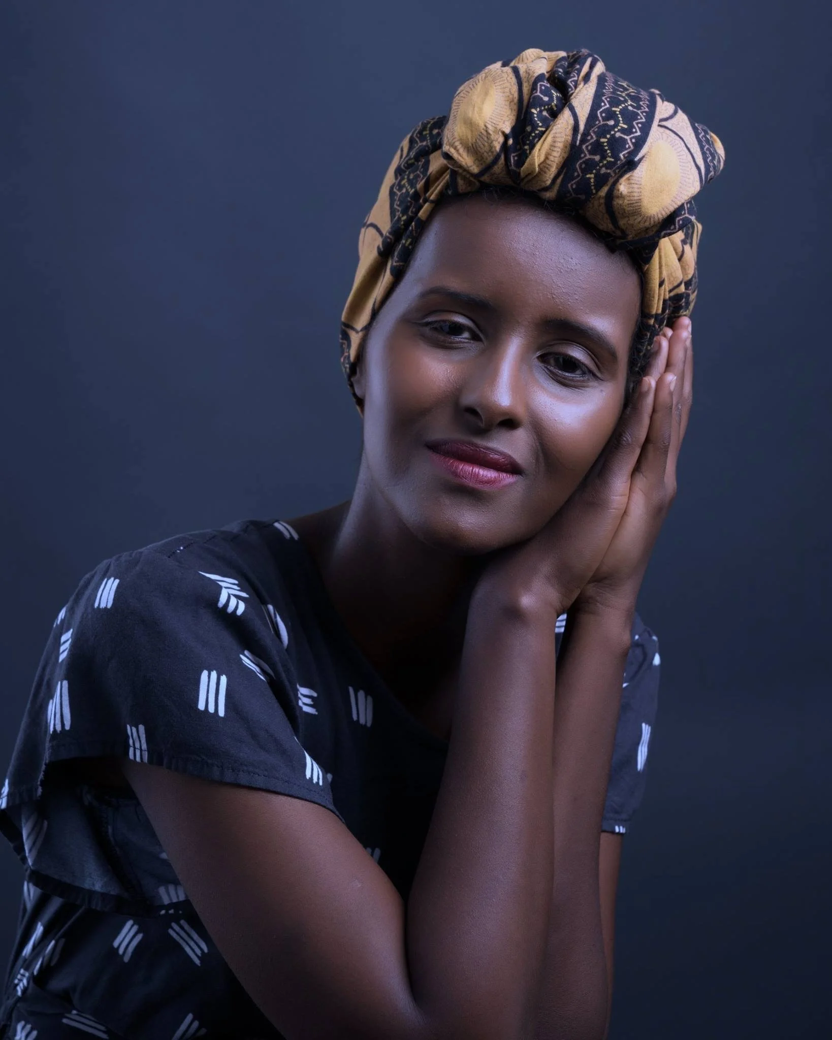 Portrait of a woman with dark skin, wearing a yellow and black patterned headwrap, resting her head on her hands with elbows on her knees, against a dark background.