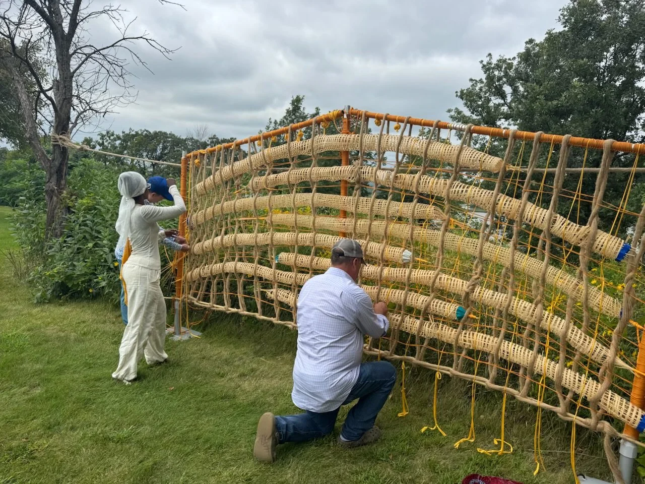 People working on a large outdoor woven bamboo structure with wooden rods and yellow ropes, on a grassy area with trees under cloudy sky.