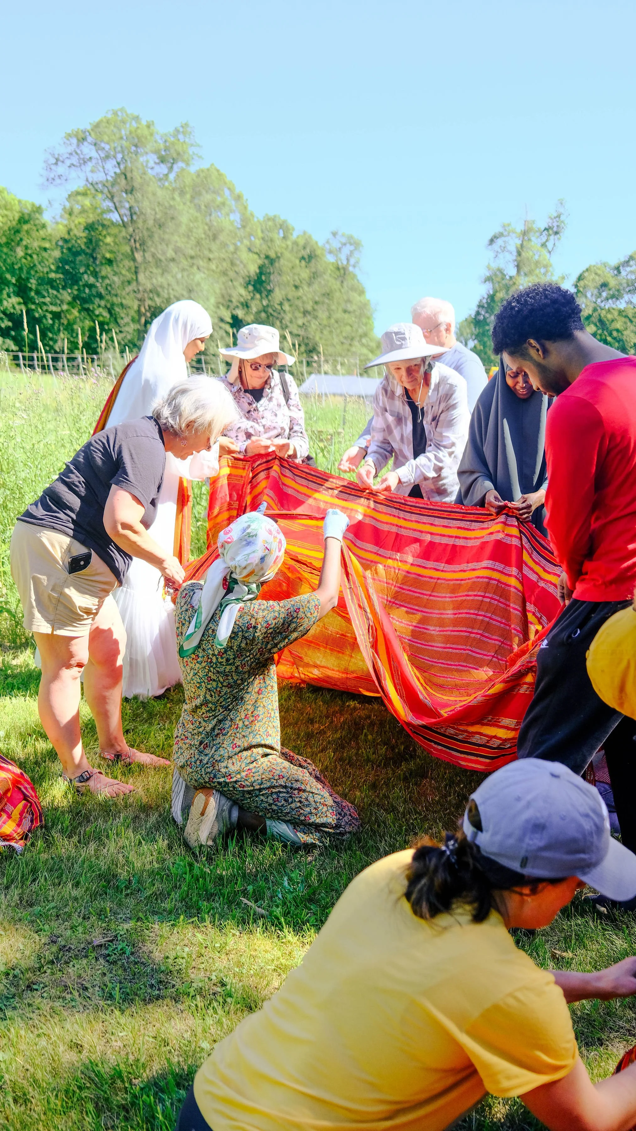 A diverse group of people working together outdoors to set up or repair a colorful fabric, with natural green trees and a blue sky in the background.