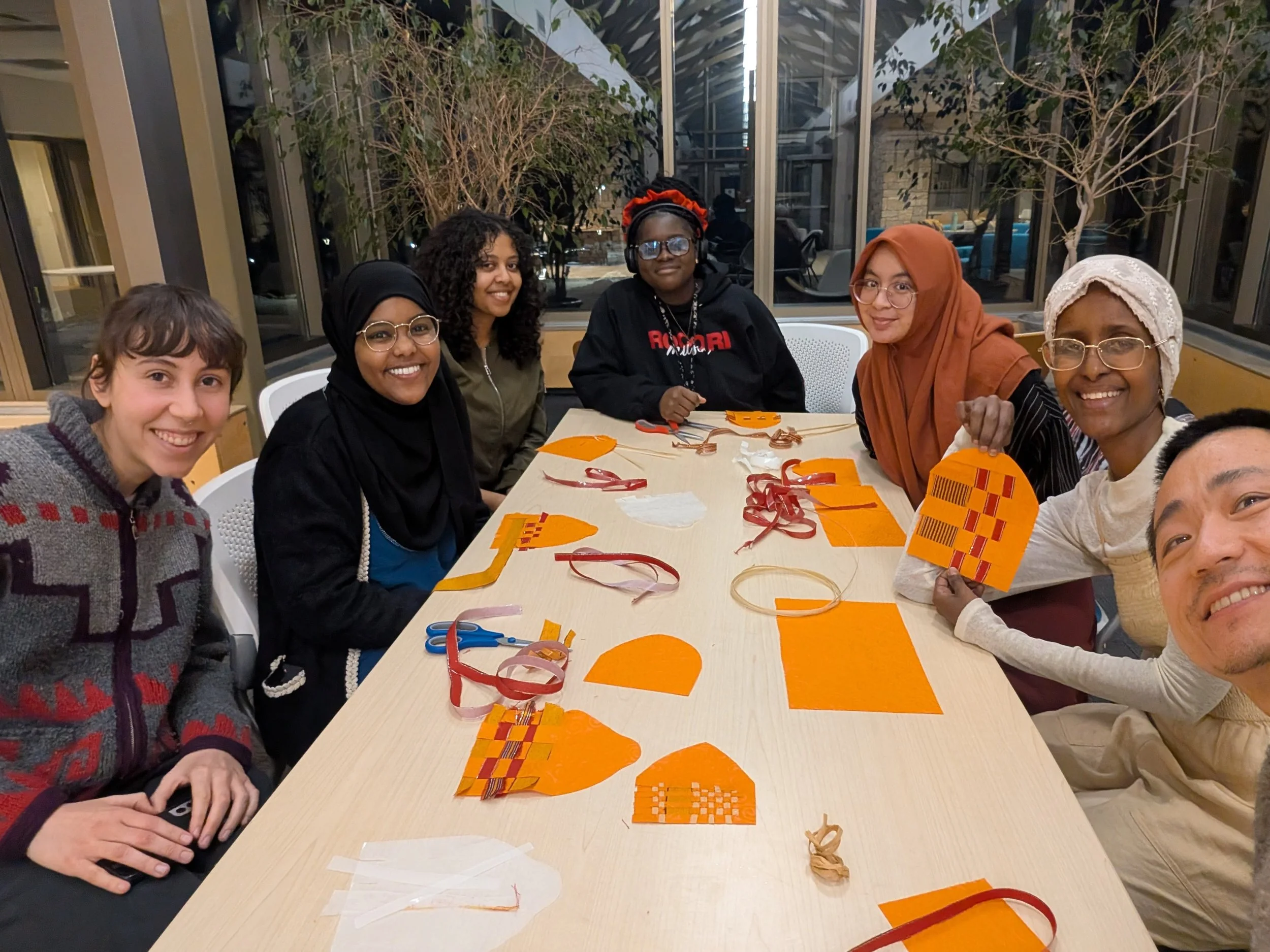 A group of six women sitting around a table at night, indoors, with orange craft supplies and scissors on the table, posing for the photo.