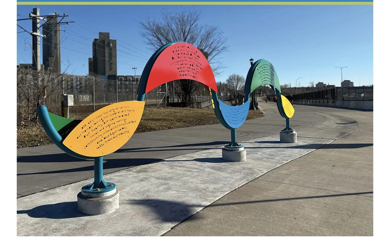 Colorful outdoor art installation of curved panels with handwritten text, each supported by a concrete base, along a paved walkway near a fence and city skyline with leafless trees under a clear blue sky.