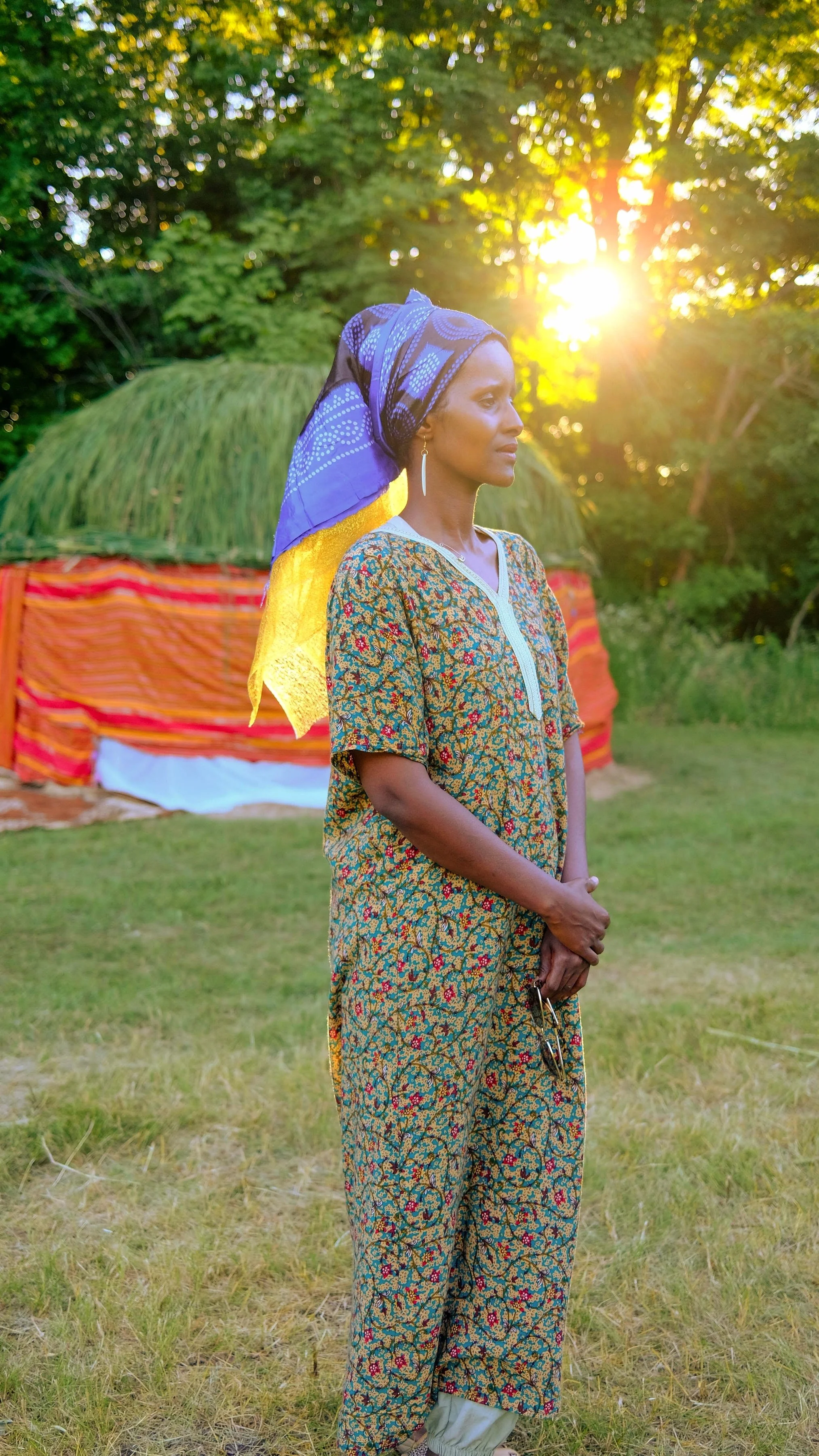A woman in colorful patterned attire standing outdoors during sunset, with a thatched hut and lush greenery in the background.