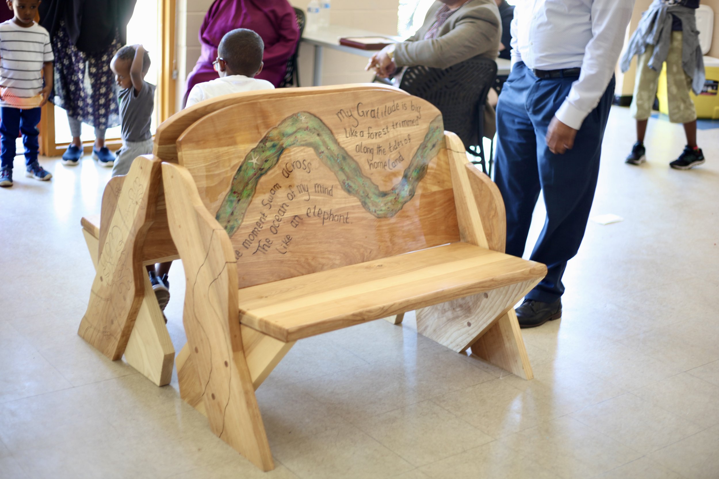 Wooden bench with painted river scene and handwritten poem, surrounded by children and adults in a room.