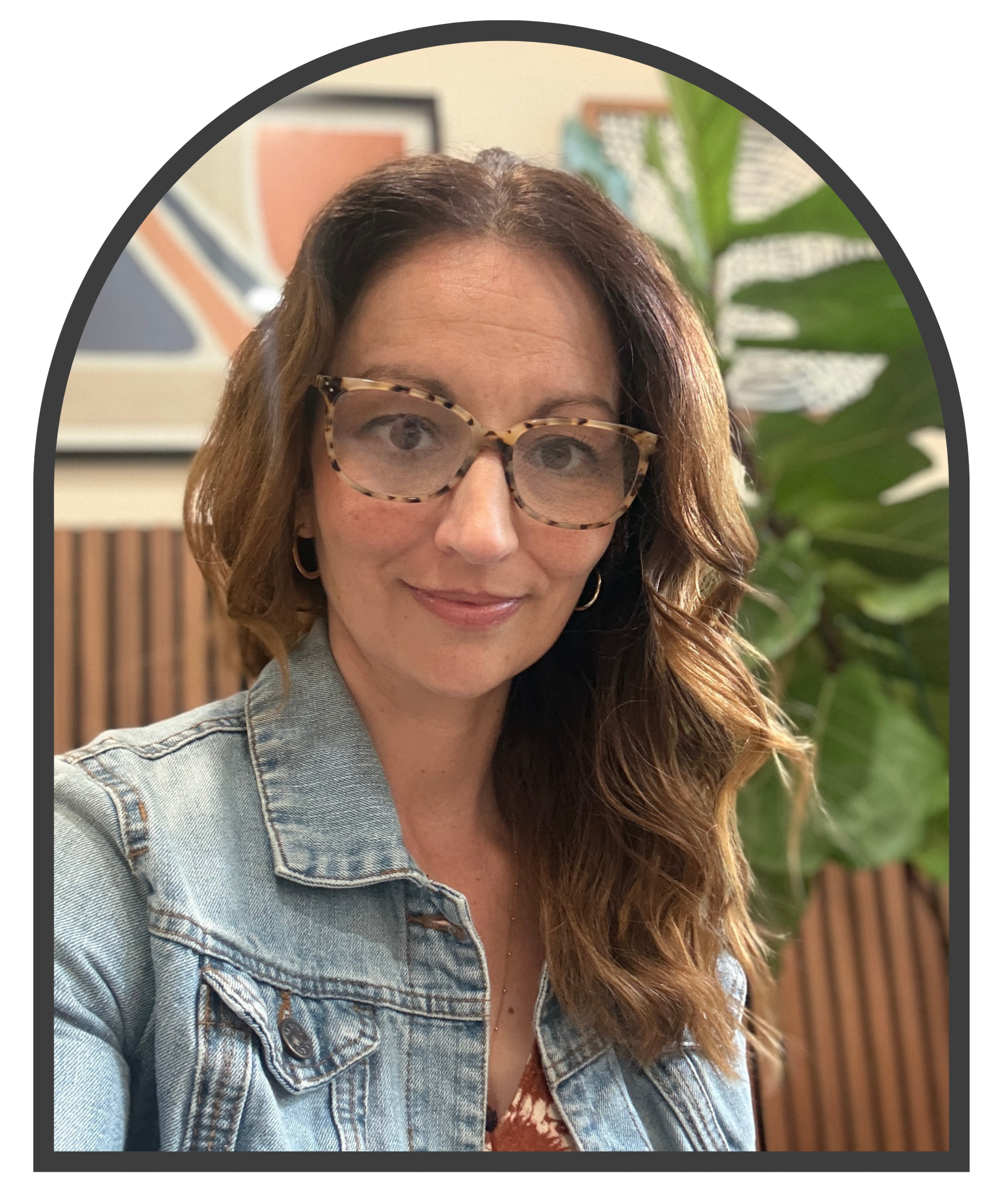 Close-up of a woman with wavy brown hair wearing patterned glasses and a denim jacket, standing indoors with art and a large green plant in the background.
