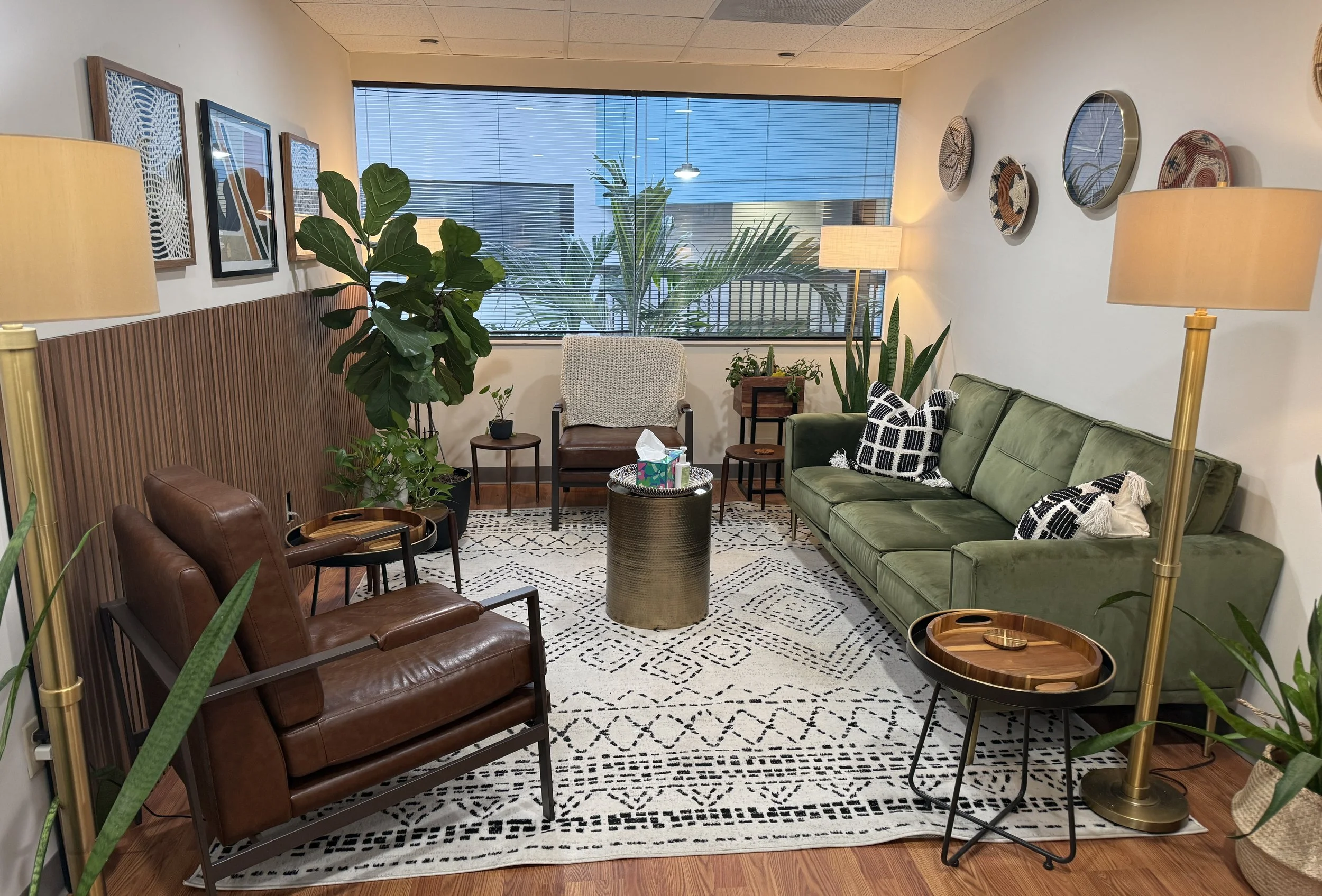 A cozy office waiting area with a green sofa, brown leather chairs, and various plants. There are two beige floor lamps, framed artwork on the wall, and decorative mirrors and baskets. Large window with blinds lets in natural light.