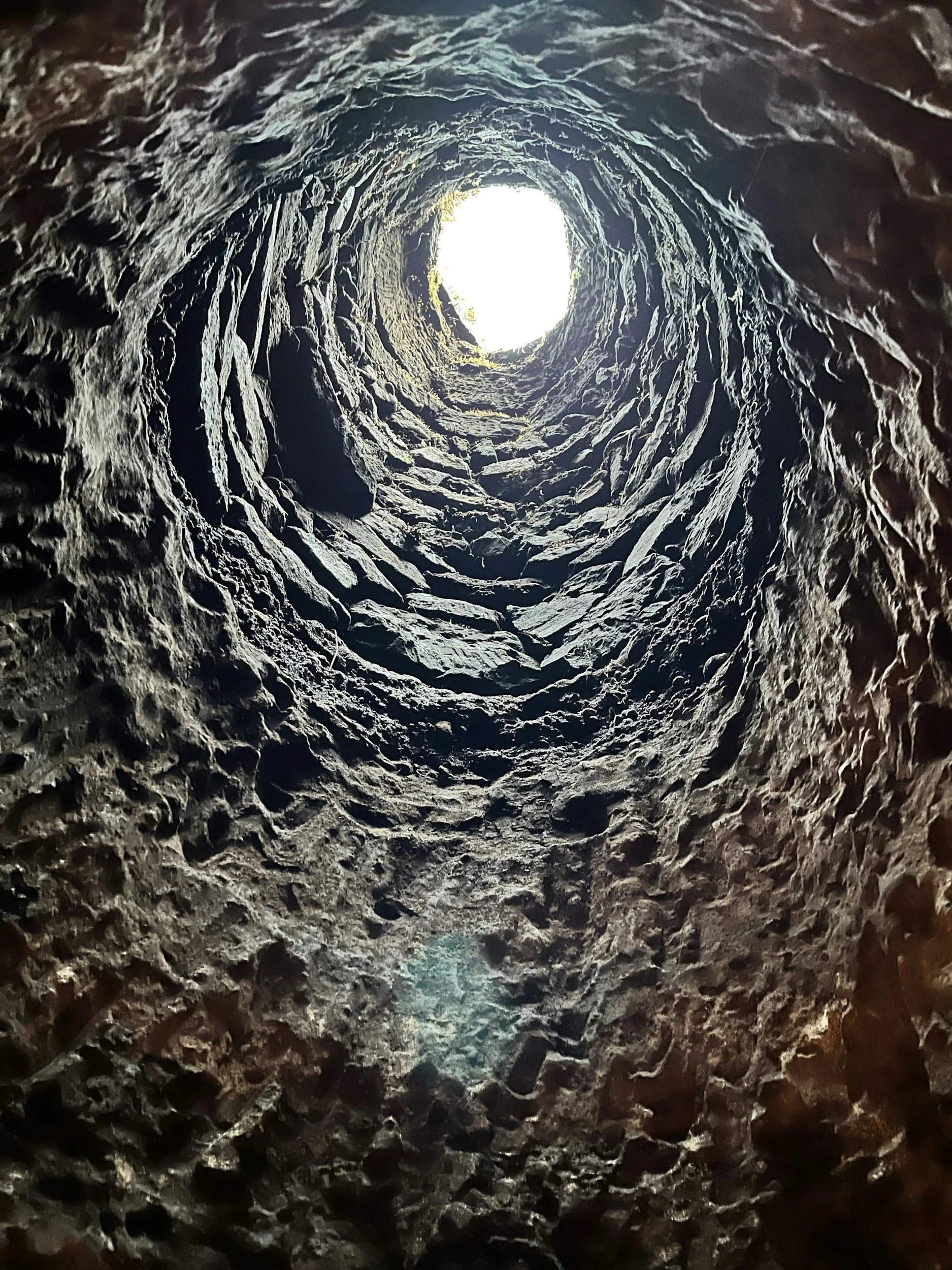 View looking up inside a deep, rough, rocky well or shaft with light at the top opening.