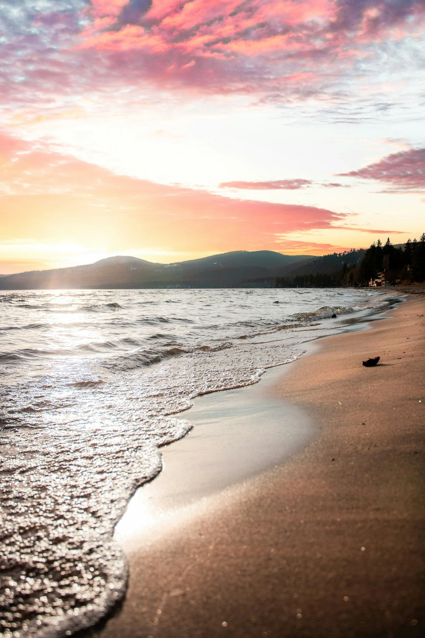 Sunset over a beach with waves gently washing onto the sandy shore, distant hills in the background, and colorful clouds in the sky.