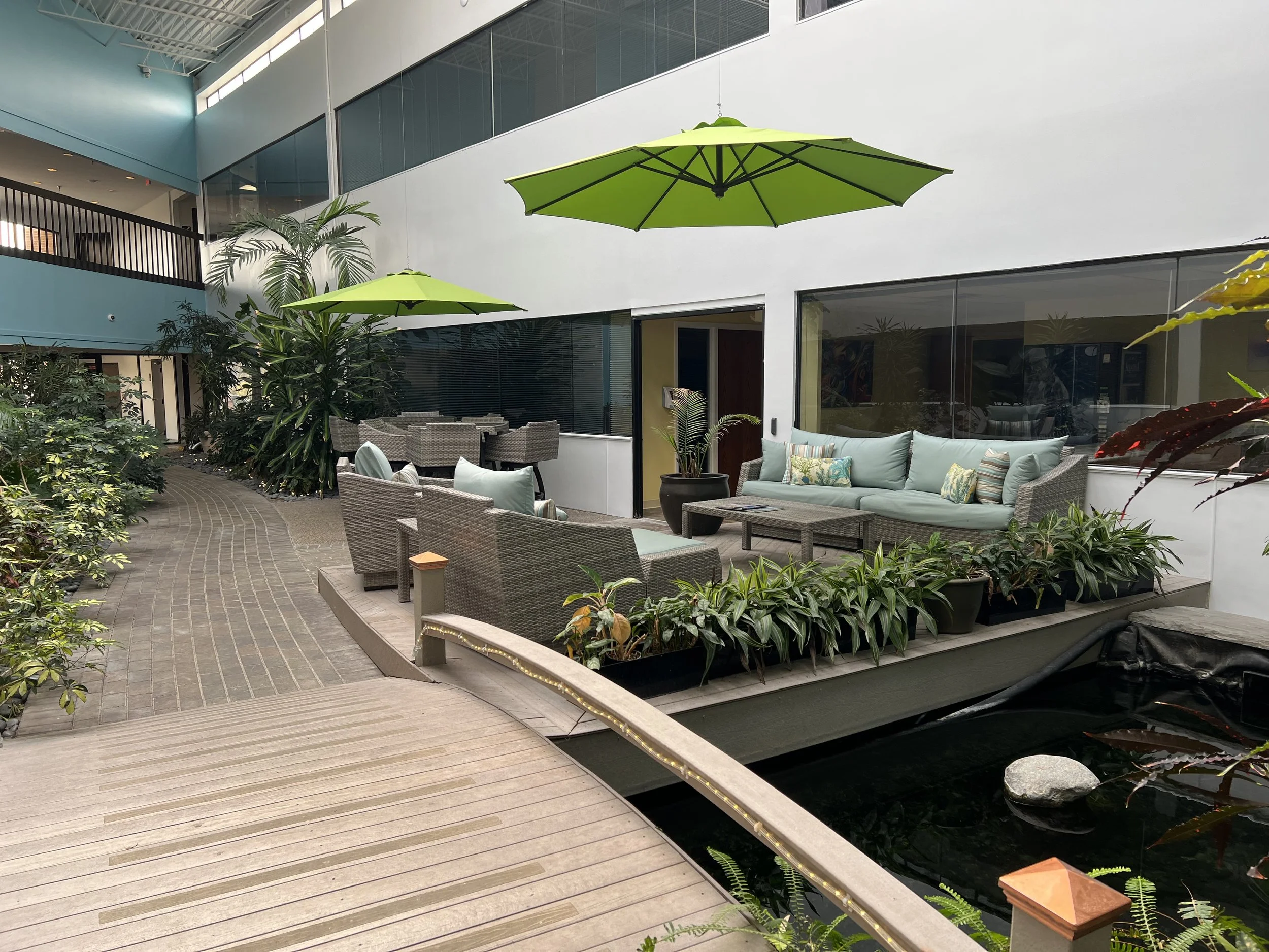 Indoor atrium with large green umbrellas, potted plants, patio furniture, glass walls, and a water feature.