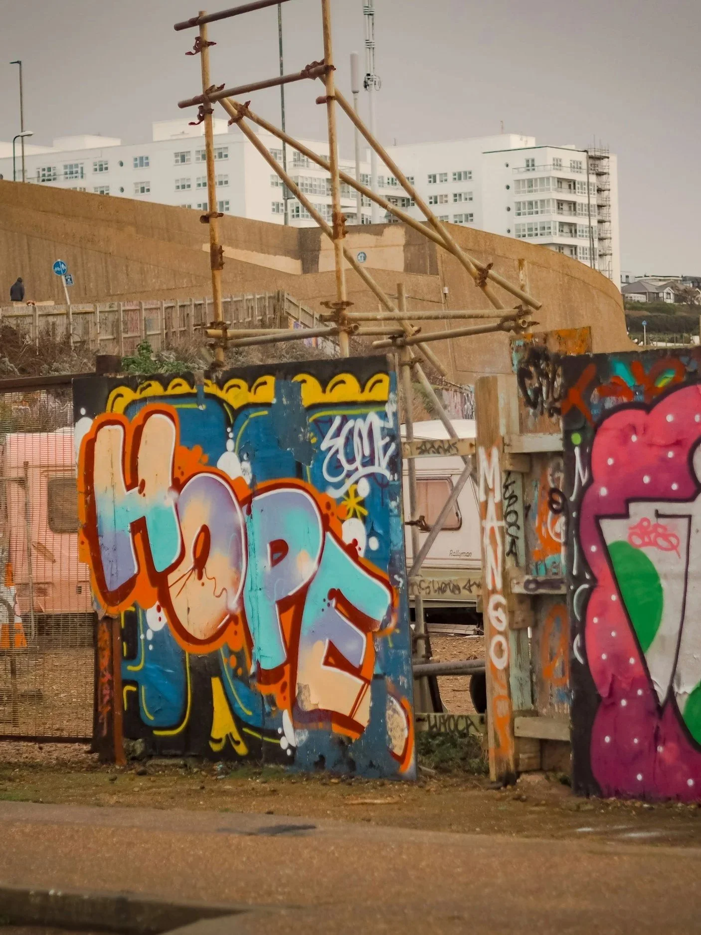 A row of colorful graffiti art on metallic panels in front of a construction site. Behind the panels, there is a tall brown construction barrier and scaffolding, with two white apartment buildings visible in the background. The sky is overcast, and there are a few street signs and a person in dark clothing walking in the distance.
