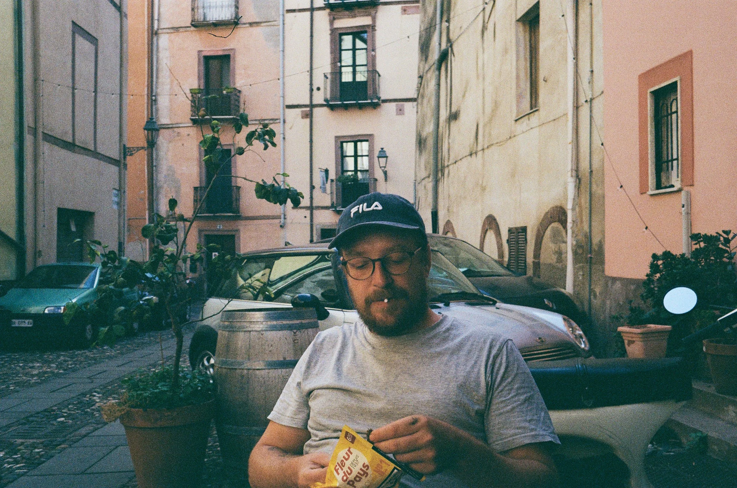 Ein Mann mit Brille, Bart und Mütze, sitzend in einer Straßen-Cafeecke, liest ein Paket und raucht.