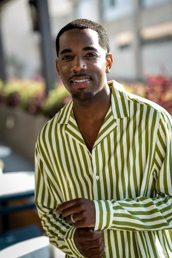 A smiling Black man wearing a green and white striped shirt, standing outdoors with blurred colorful plants and buildings in the background.