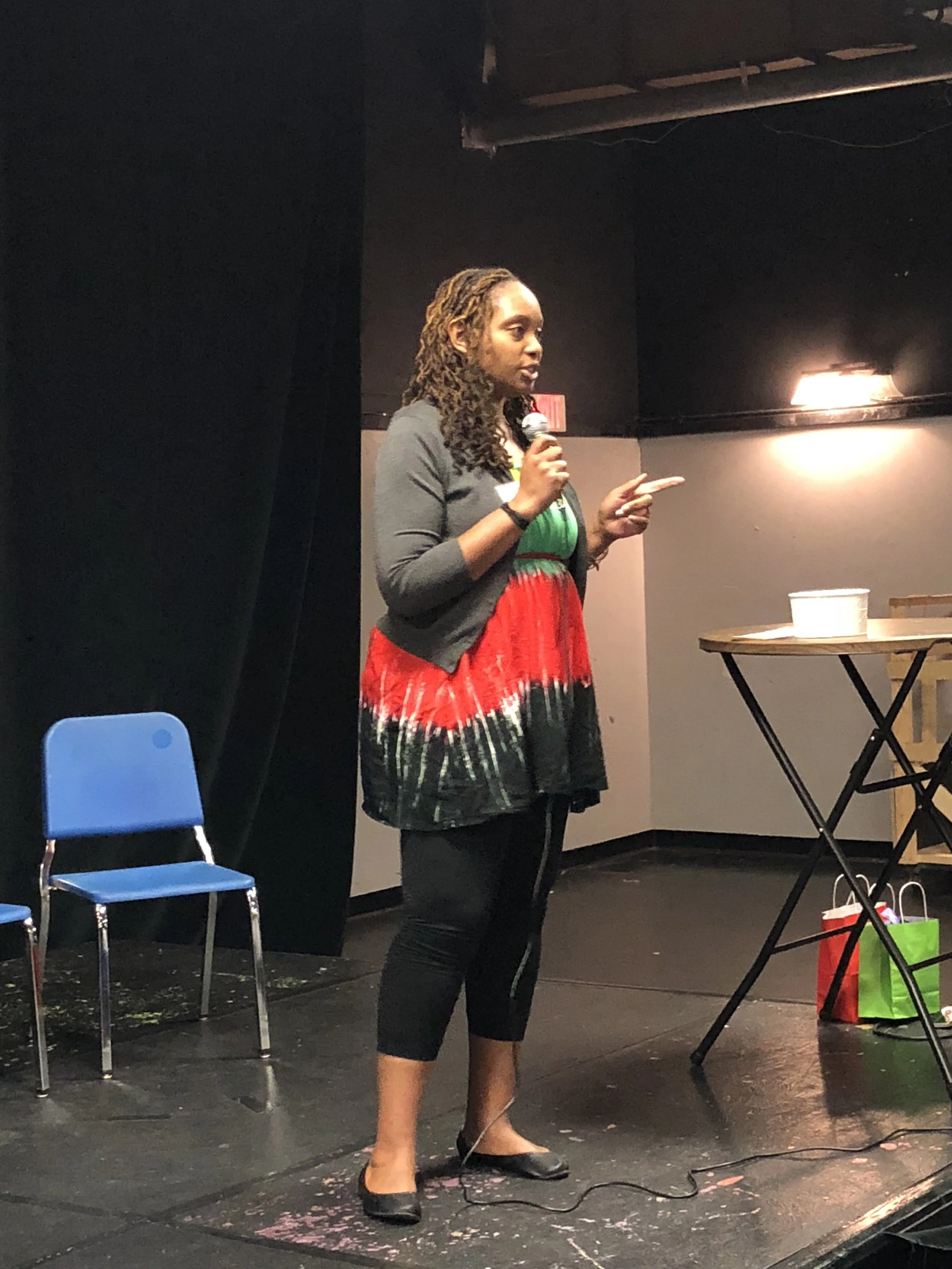 A woman with curly hair stands on a stage, holding a microphone and speaking. She is wearing a colorful dress with red, green, and black patterns, paired with a gray cardigan, and black shoes. There are two empty blue chairs to her left, a table with a paper cup, and gift bags on the right.