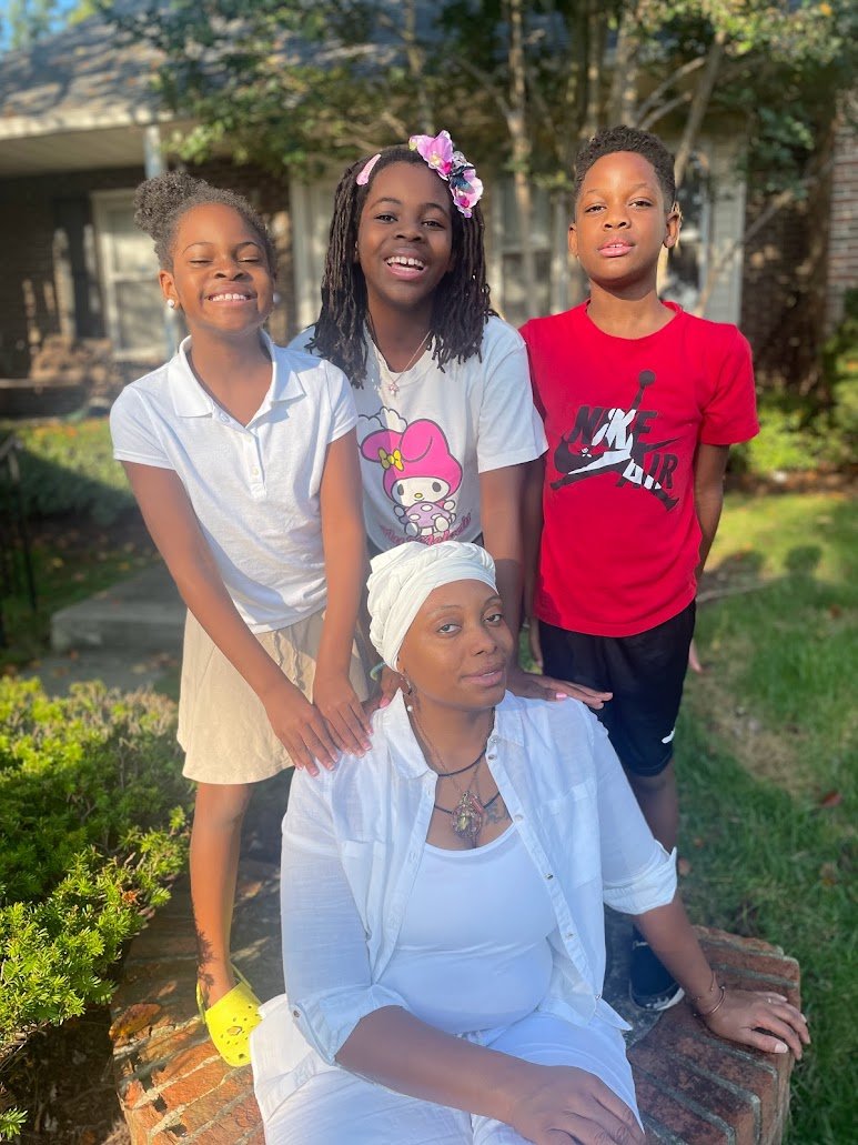 Four children and one woman outdoors, with greenery and a house in the background. The woman is seated on a brick bench wearing a white headwrap and white clothing. The children are standing around her, smiling, with two girls and a boy. The girl on the left has pigtails and a white shirt. The girl in the middle has dreadlocks, a floral headband, and a graphic T-shirt. The boy on the right has short hair and is wearing a red Nike Air T-shirt.