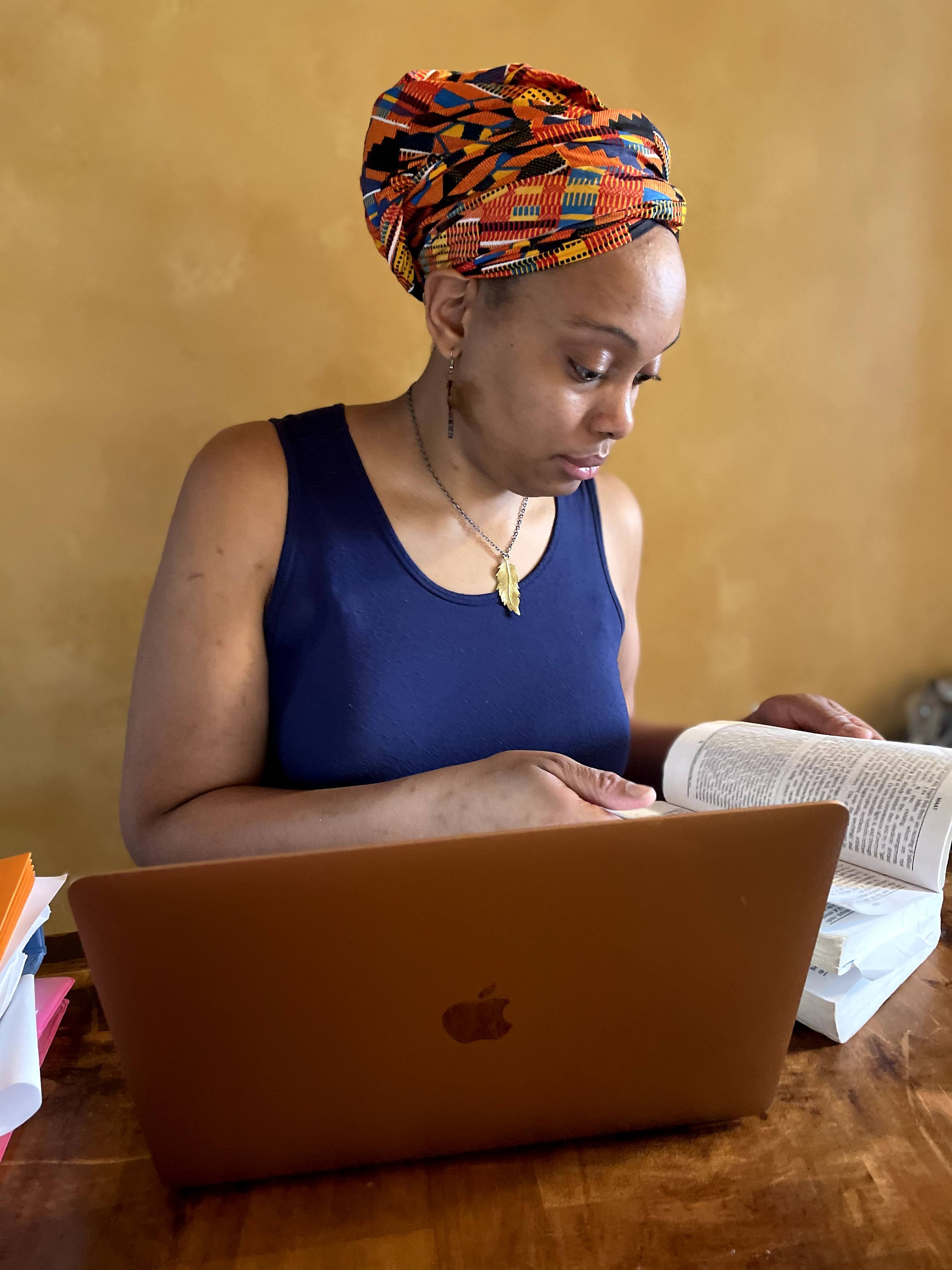 A woman with a colorful headwrap, wearing a navy dress, sitting at a wooden table, looking at a book while working on a laptop.