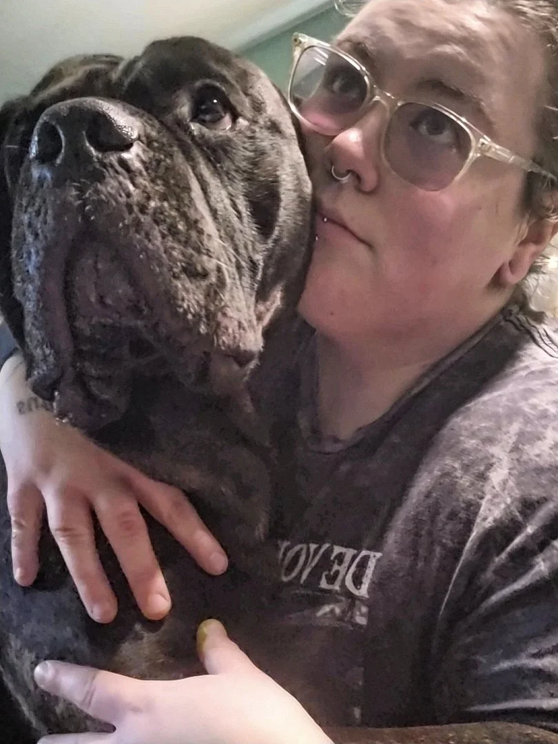 A woman with glasses and piercings hugging a large brindle dog.