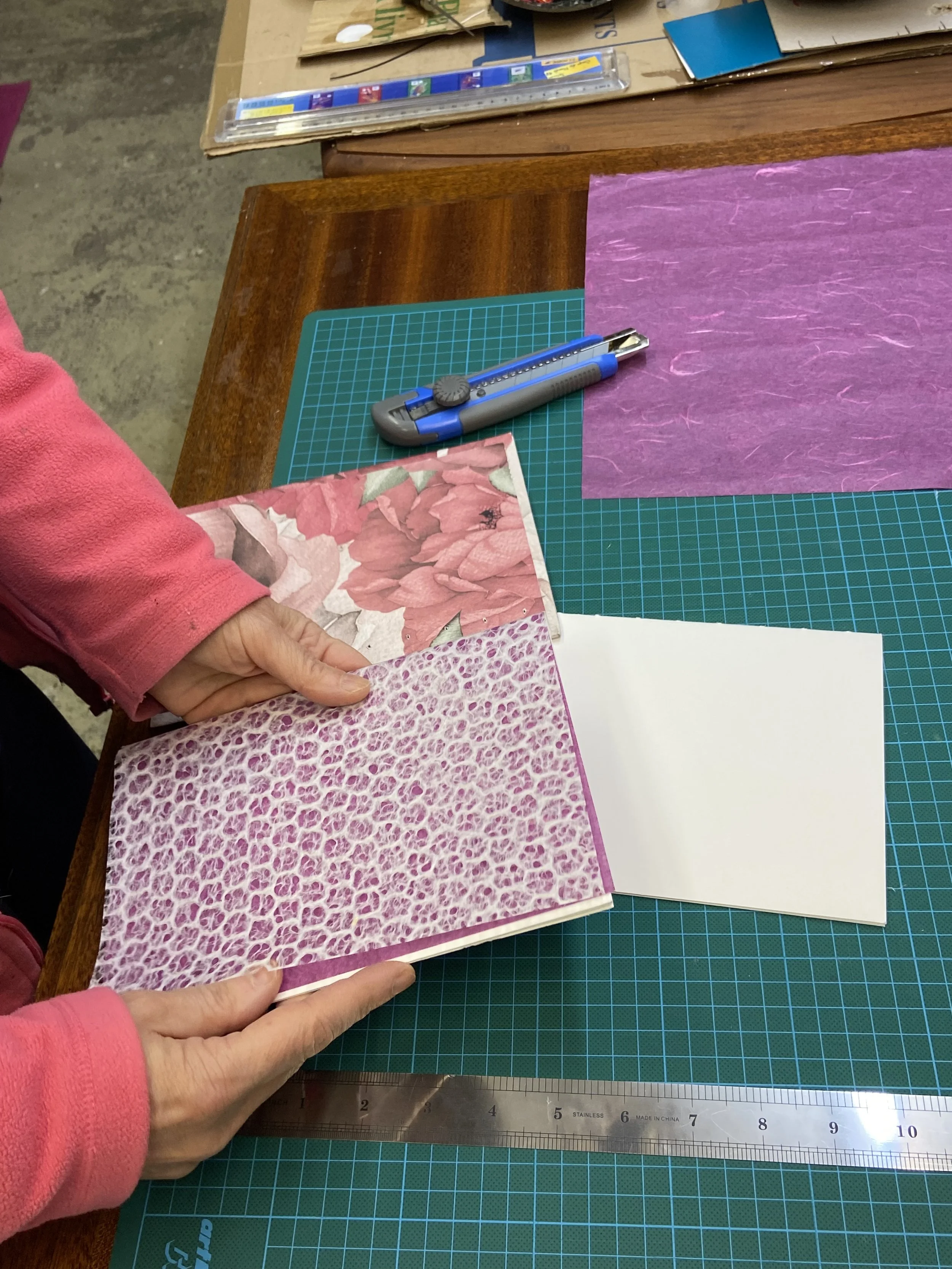 Une personne tient un livre avec une couverture de motifs floraux rose et un motif animalier rose et blanc, posé sur une table avec un tapis de découpe vert, un cutter et des feuilles de papier coloré.