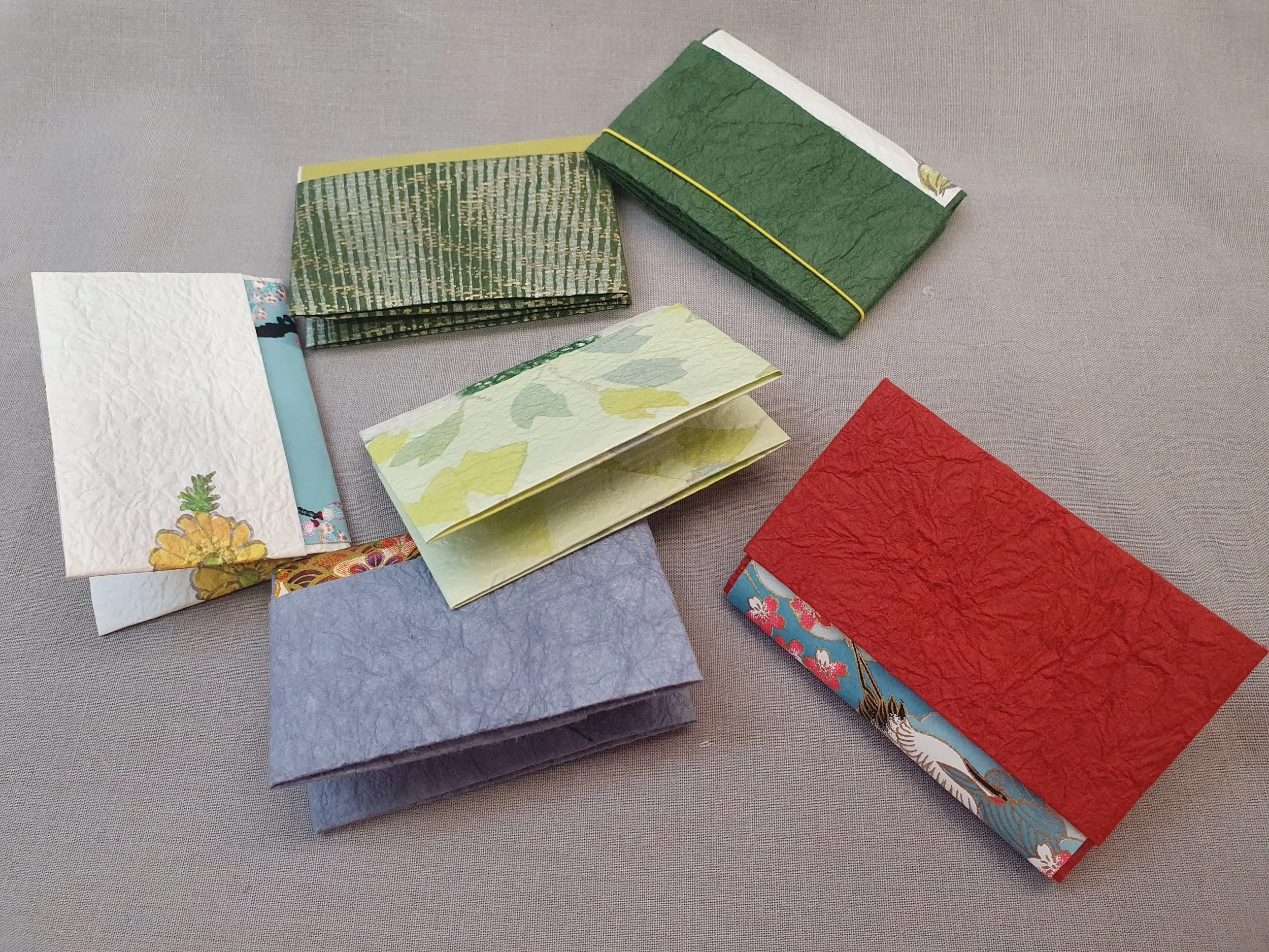 Plusieurs petits livres ou carnets colorés en papier fabriqués à la main, disposés sur une surface grise.