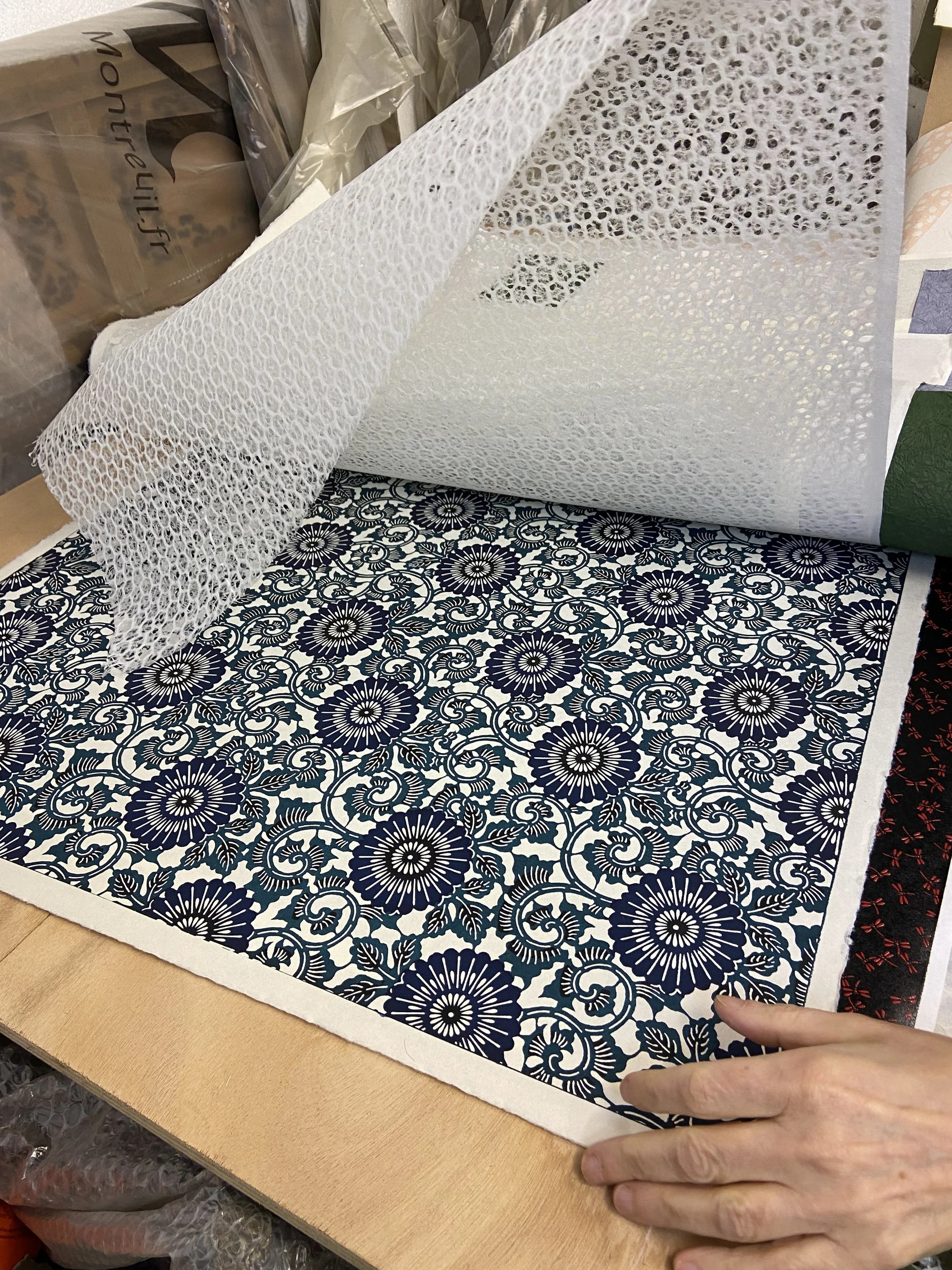Une personne déployant un papier peint à motif floral bleu et blanc sur une surface en bois, avec d'autres rouleaux de papier peint autour.