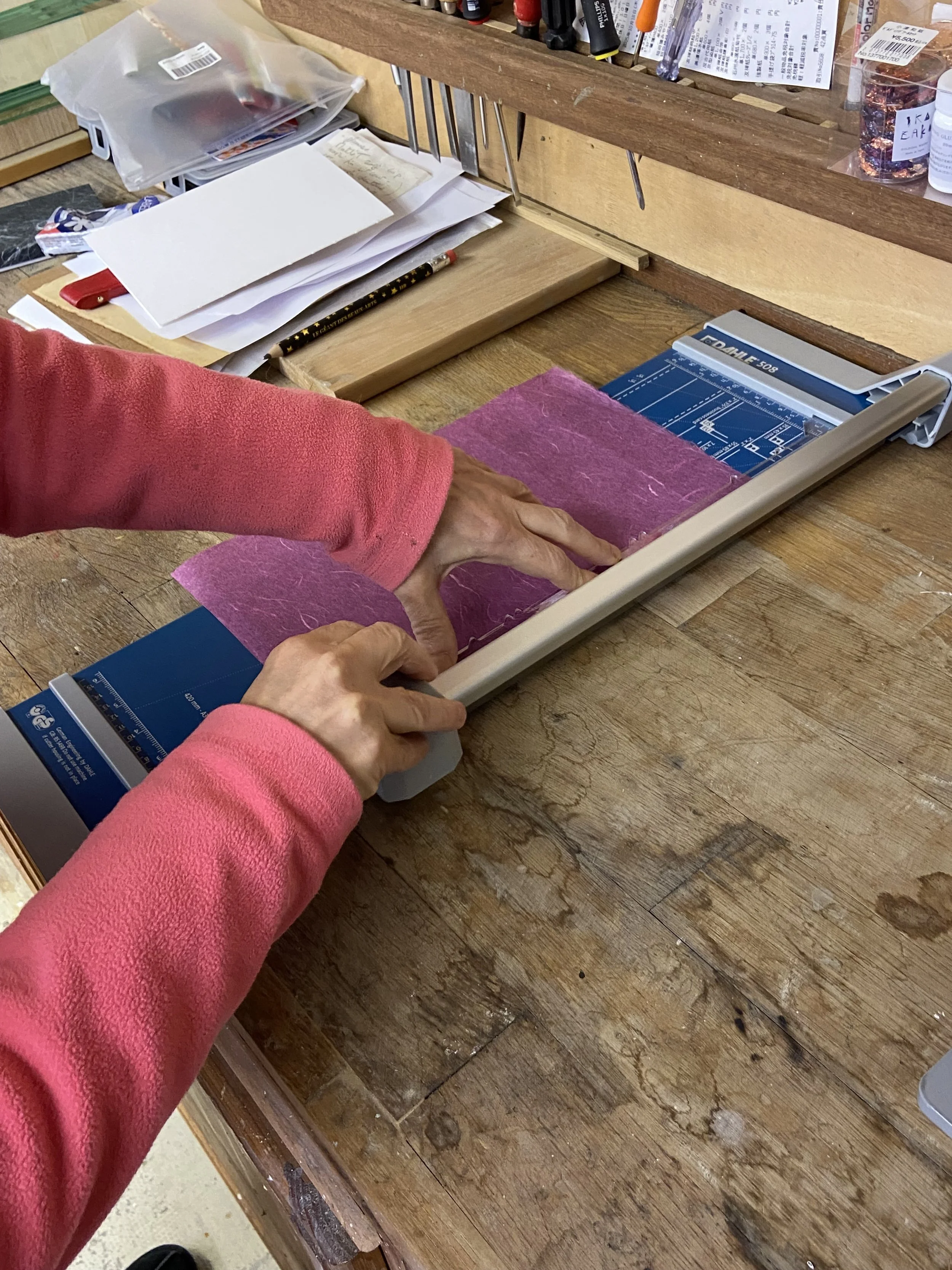 Une personne utilise une machine de découpe de papier ou de tissu sur un plan de travail en bois dans un atelier. Le plan de travail est encombré de papiers, de fournitures et d'outils.